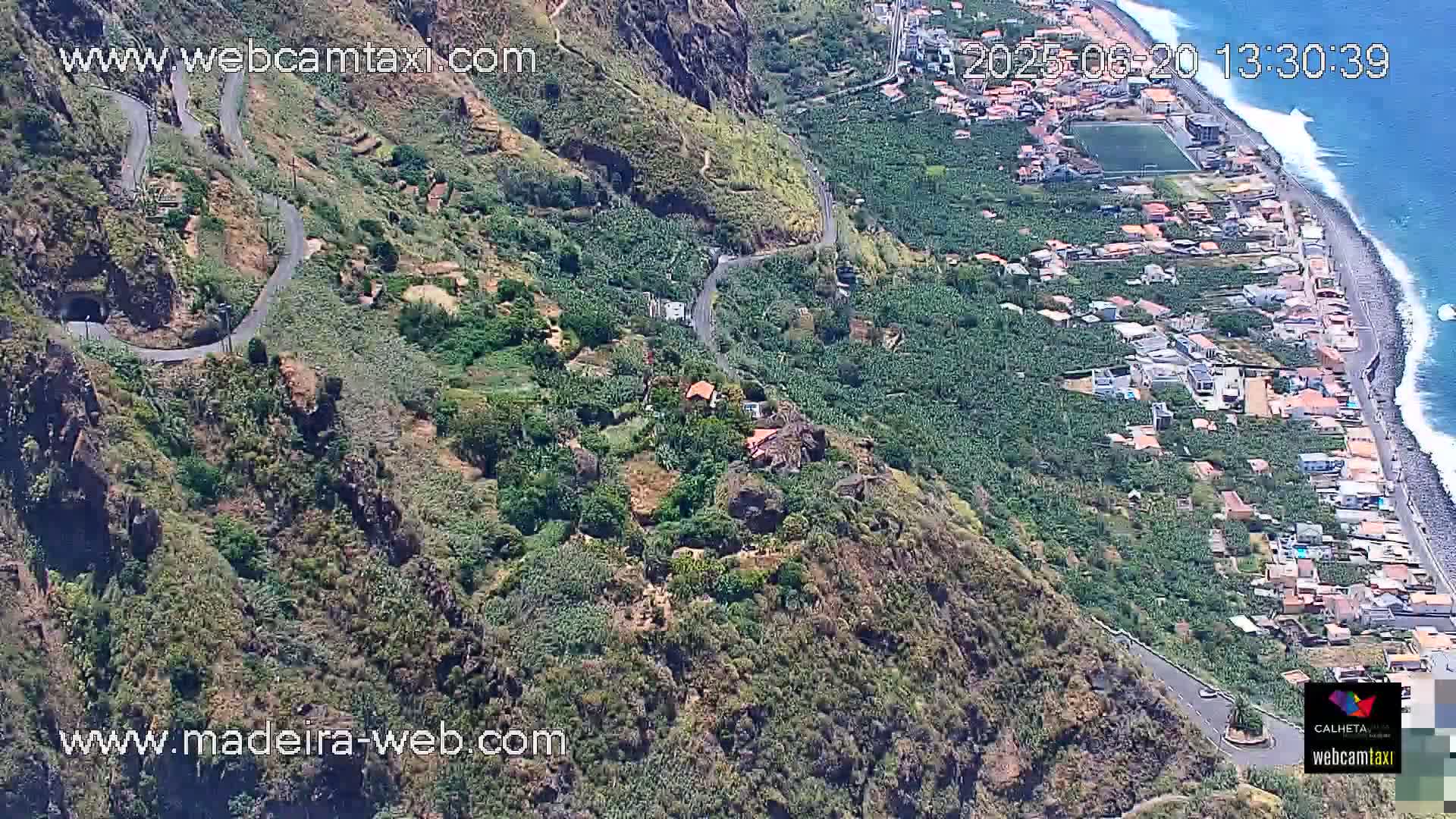 A winding road descends a steep, green hillside overlooking a coastal town with a rocky beach and ocean under sunny skies.