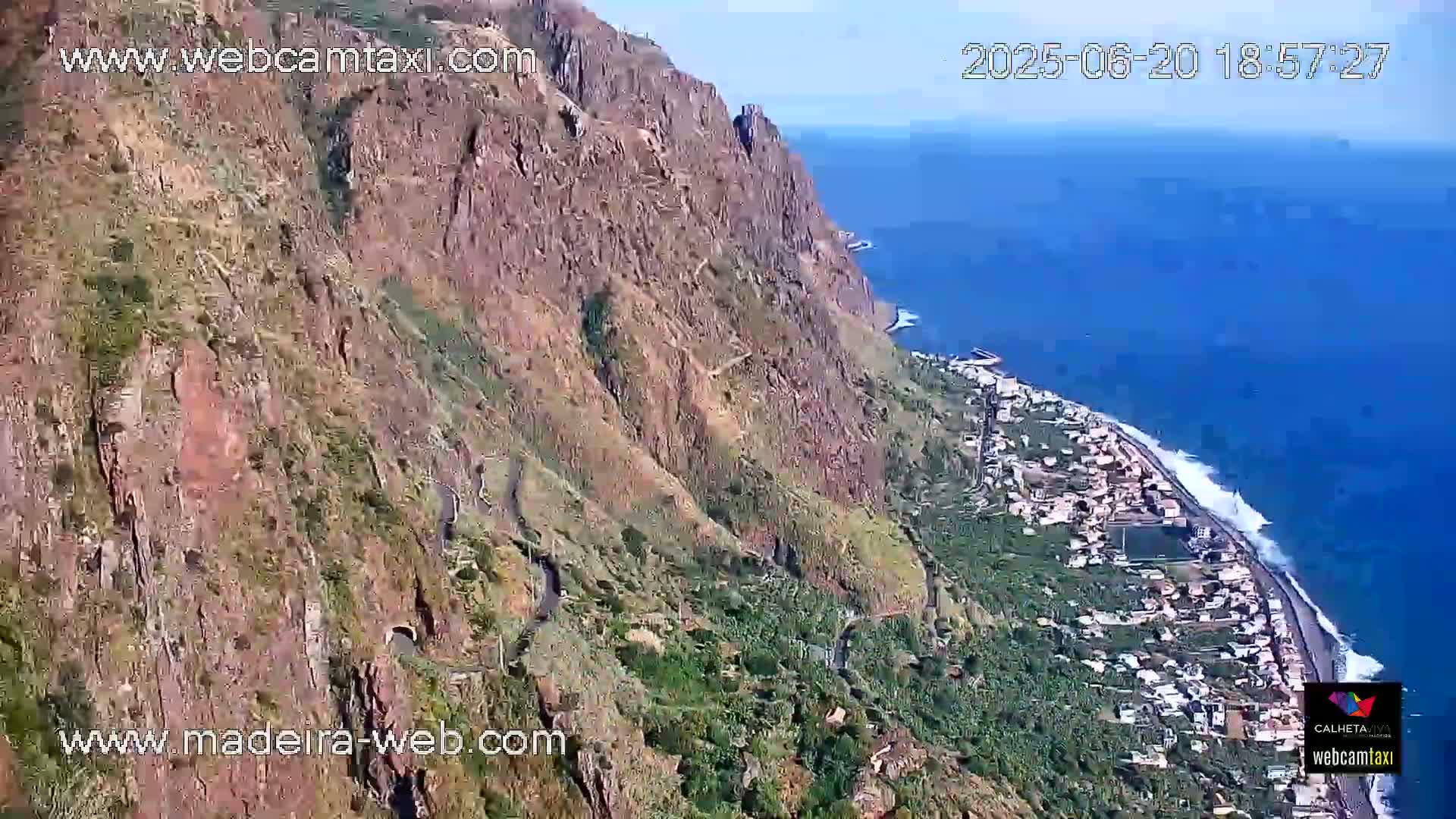 A high-angle view shows a steep, rocky cliff overlooking a coastal town and ocean with waves crashing on the shore under a clear sky.