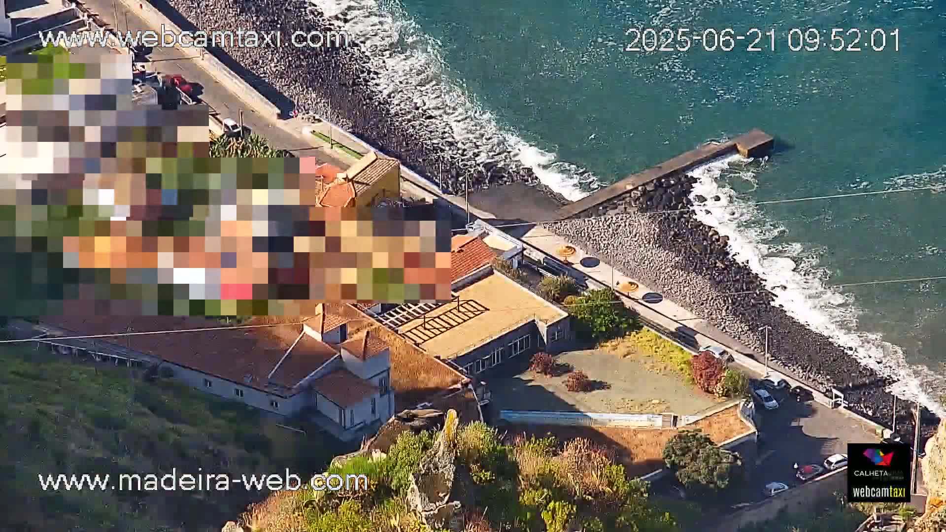An aerial view shows a rocky coastline with a building complex near the ocean under sunny conditions.