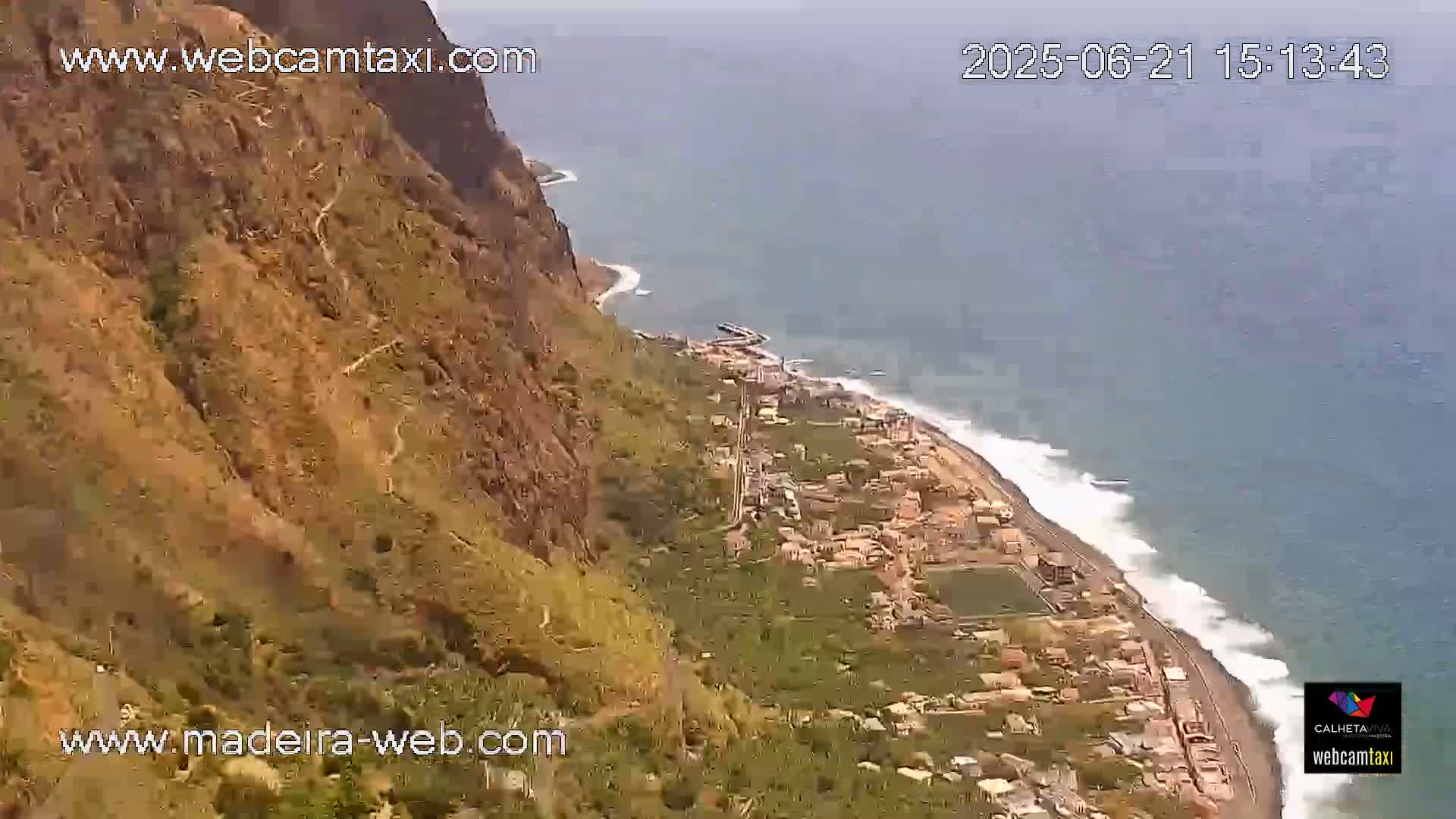 A high-angle view shows a coastal town nestled between a steep, rocky cliff and the ocean, where waves break along a sandy beach under sunny skies.
