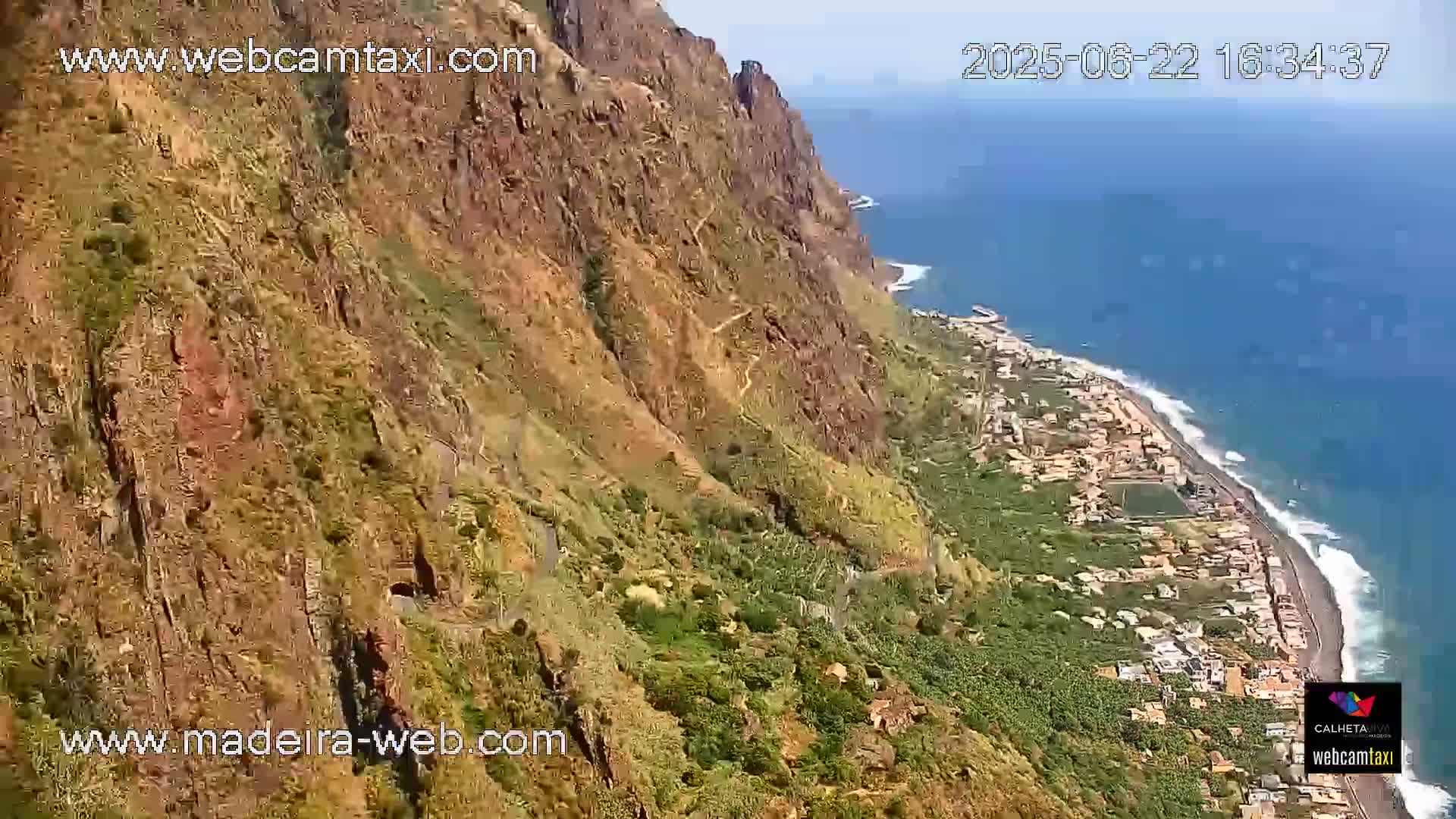 A high-angle view shows a steep, rocky cliff overlooking a coastal town and ocean on a sunny day.