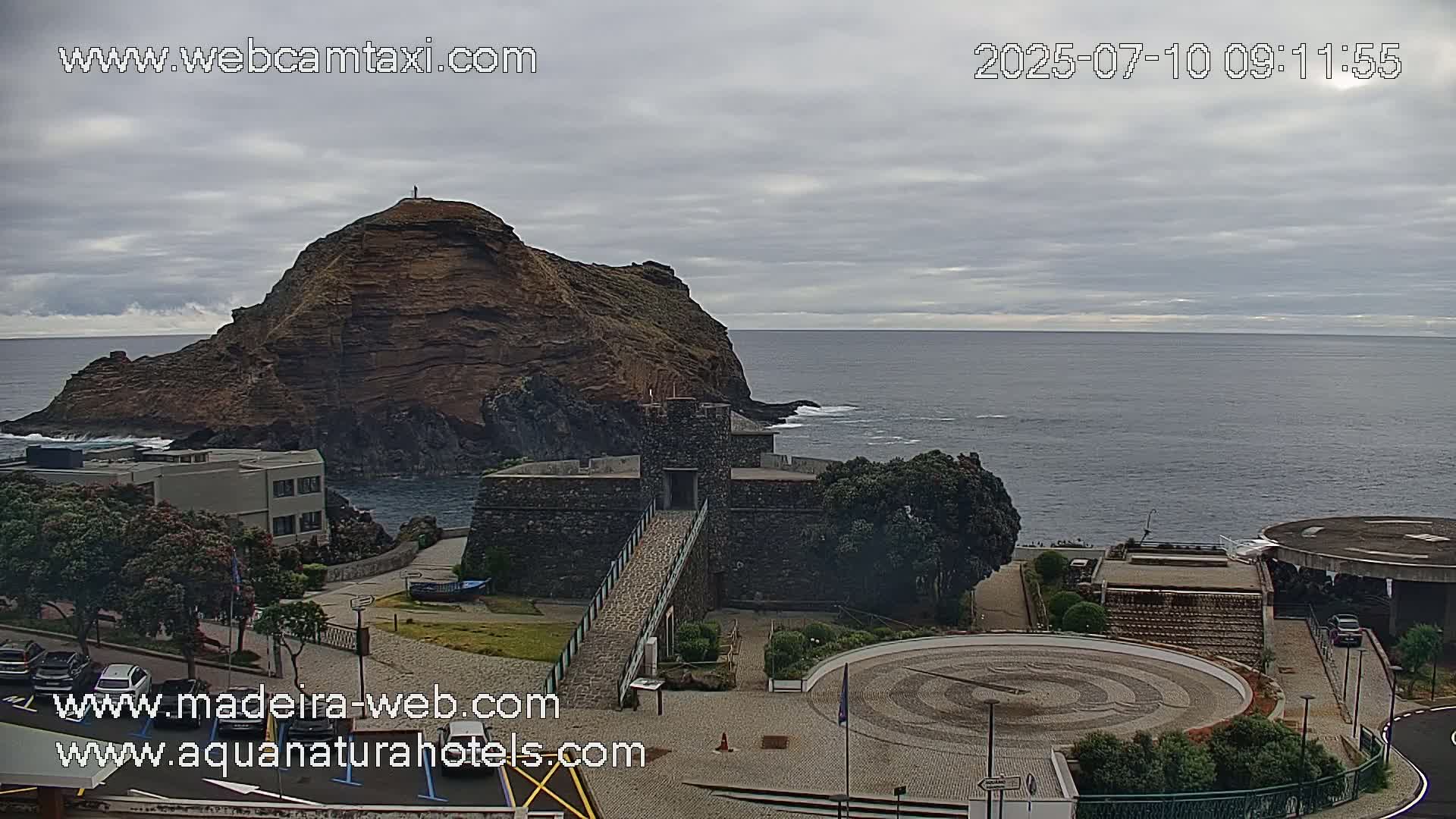A rocky island sits off a coastline, next to a stone structure with a walkway, under an overcast sky.