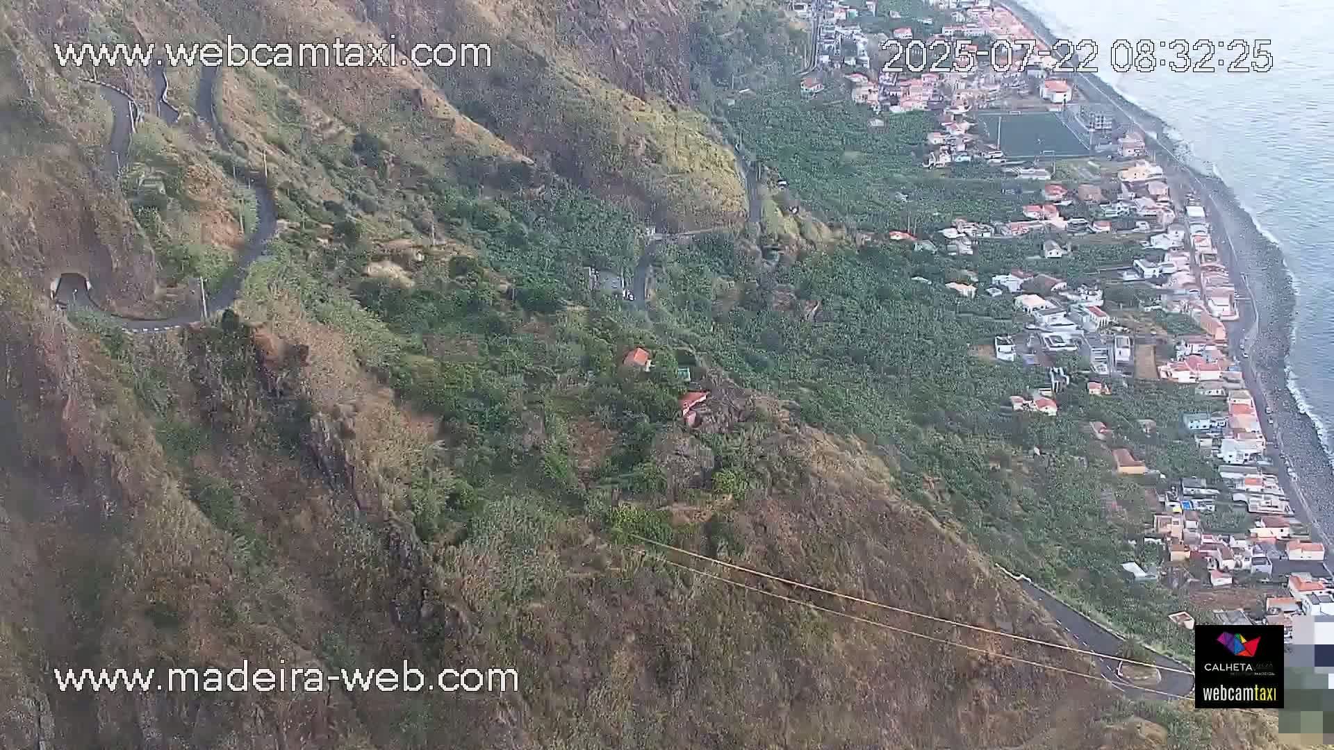 A winding road snakes down a steep, verdant hillside toward a coastal town and ocean.