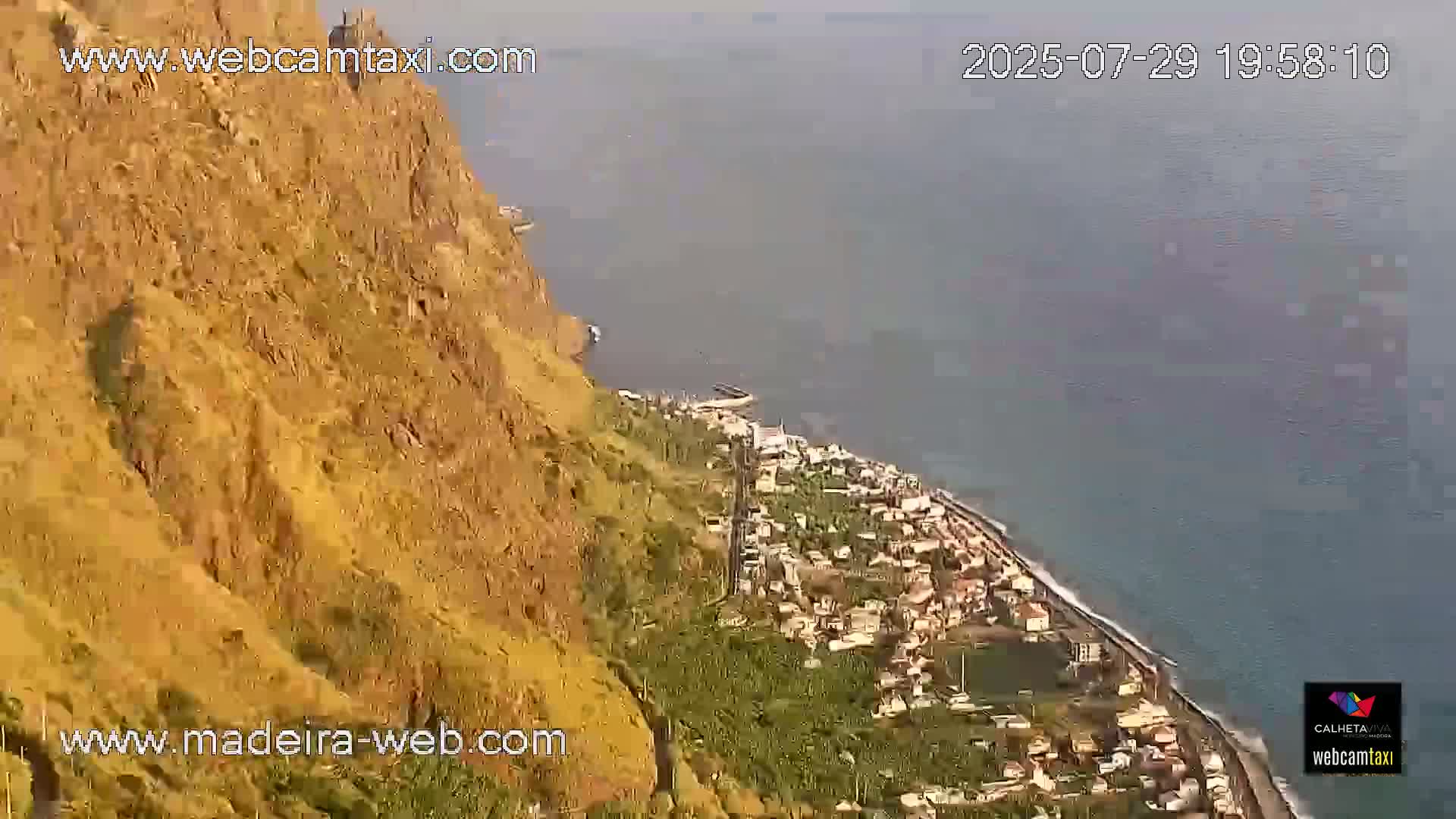 A winding road snakes down a steep, verdant hillside toward a coastal town and ocean.