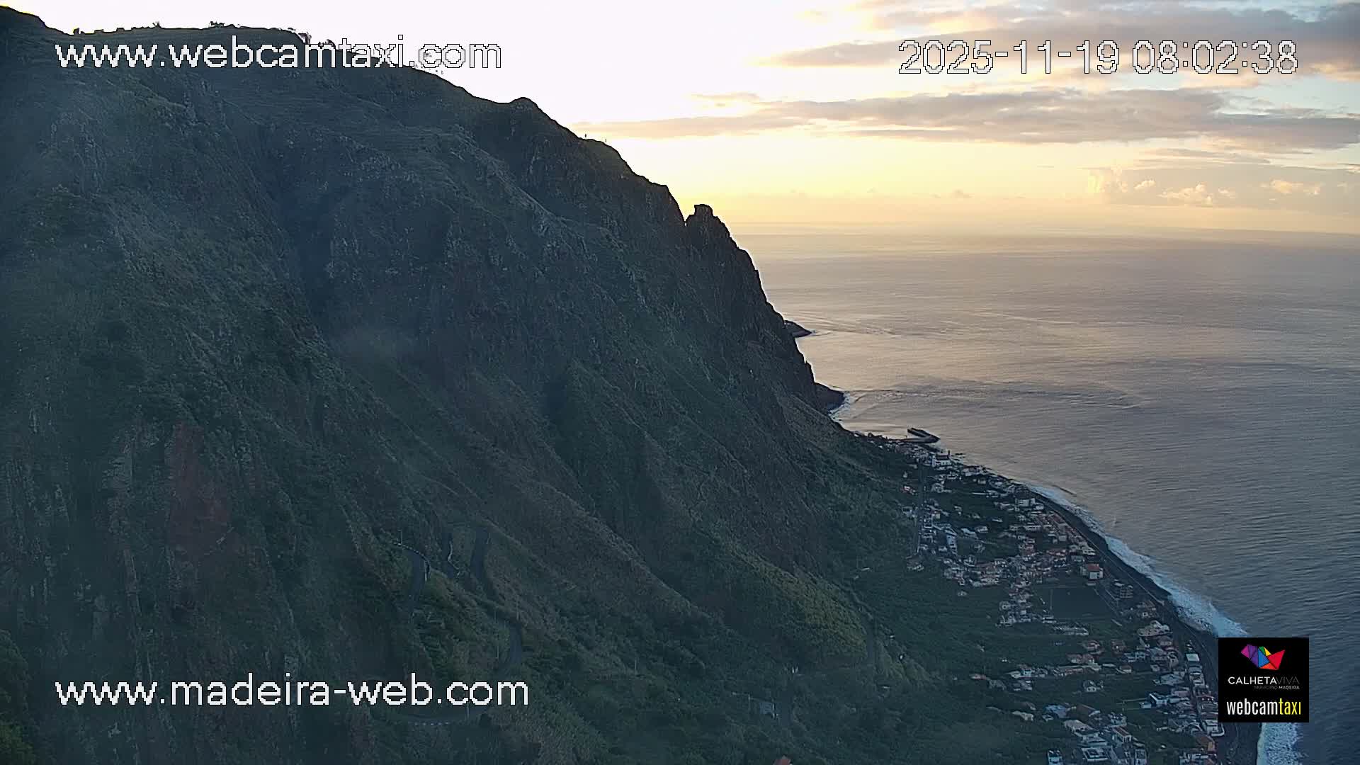Steep, verdant mountains cascade down to a coastal town nestled along a calm ocean, all bathed in the golden light of a clear sunrise with scattered clouds.