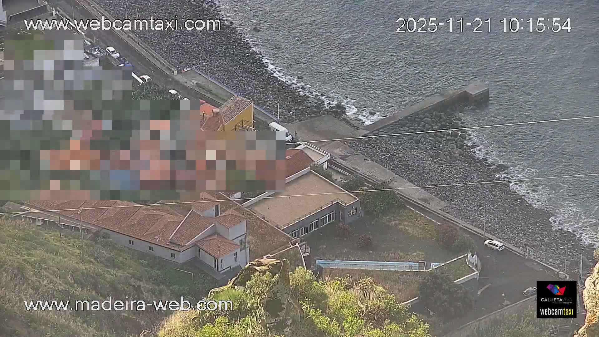 The image captures a clear and sunny day over a coastal area, featuring a mix of buildings on a hillside, a road running alongside a rocky beach, and the ocean with a concrete jetty extending into the water.