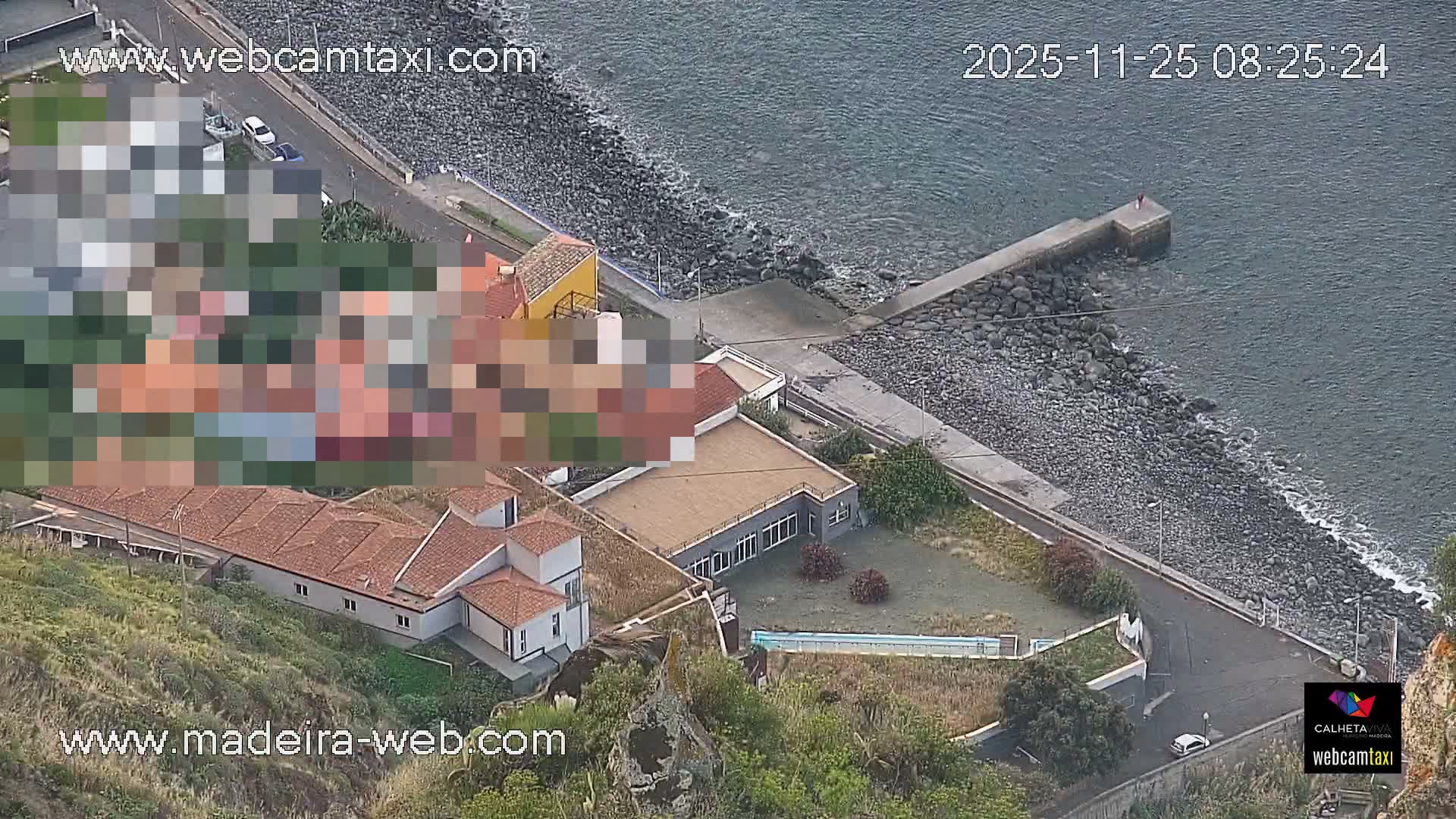 The image displays an elevated view of a tranquil coastal scene featuring a rocky shoreline, a small concrete pier extending into calm waters, a road with a few vehicles, and a scattering of buildings including one with an outdoor pool, all under bright daylight conditions.