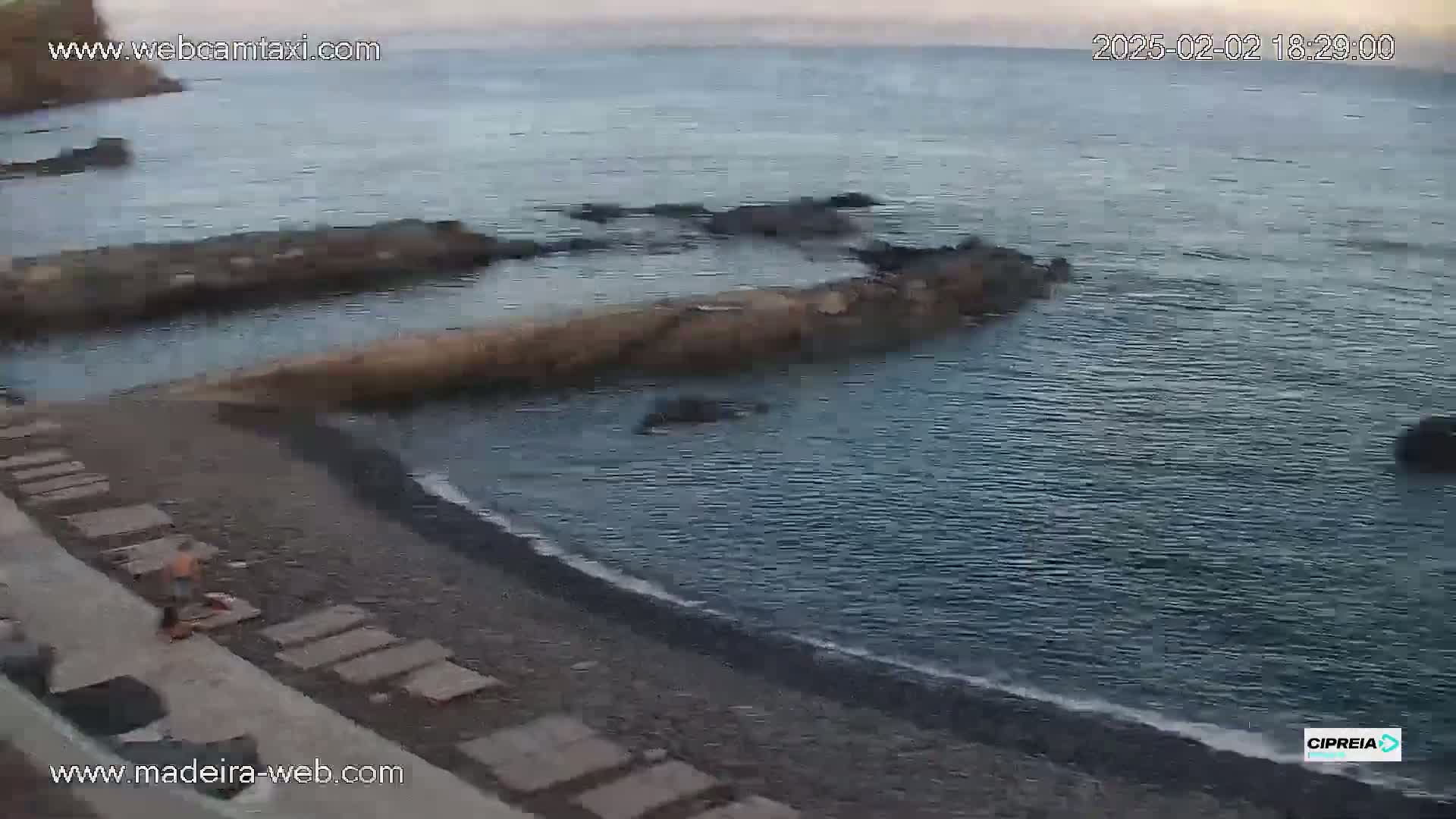 A dark-colored beach with a few people on it slopes gently down to a calm, dark-blue sea dotted with dark rocks.