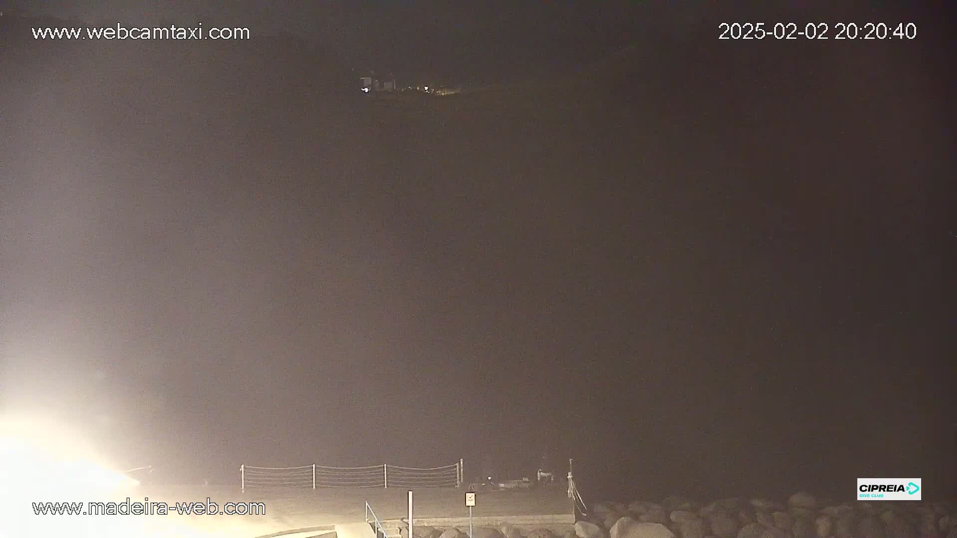 A nighttime, foggy view of a shoreline with a low fence and some rocks in the foreground.