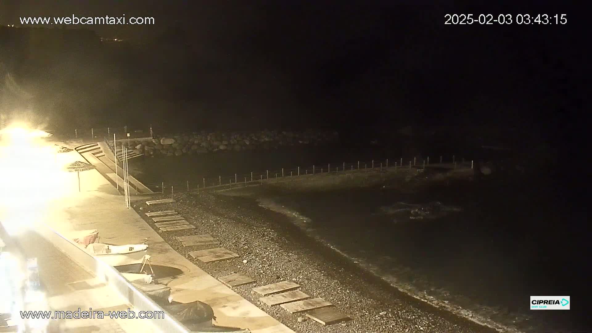 A dark, nighttime beach scene shows a pebble shoreline, calm water lapping at the shore, and a low wall separating the beach from the water.