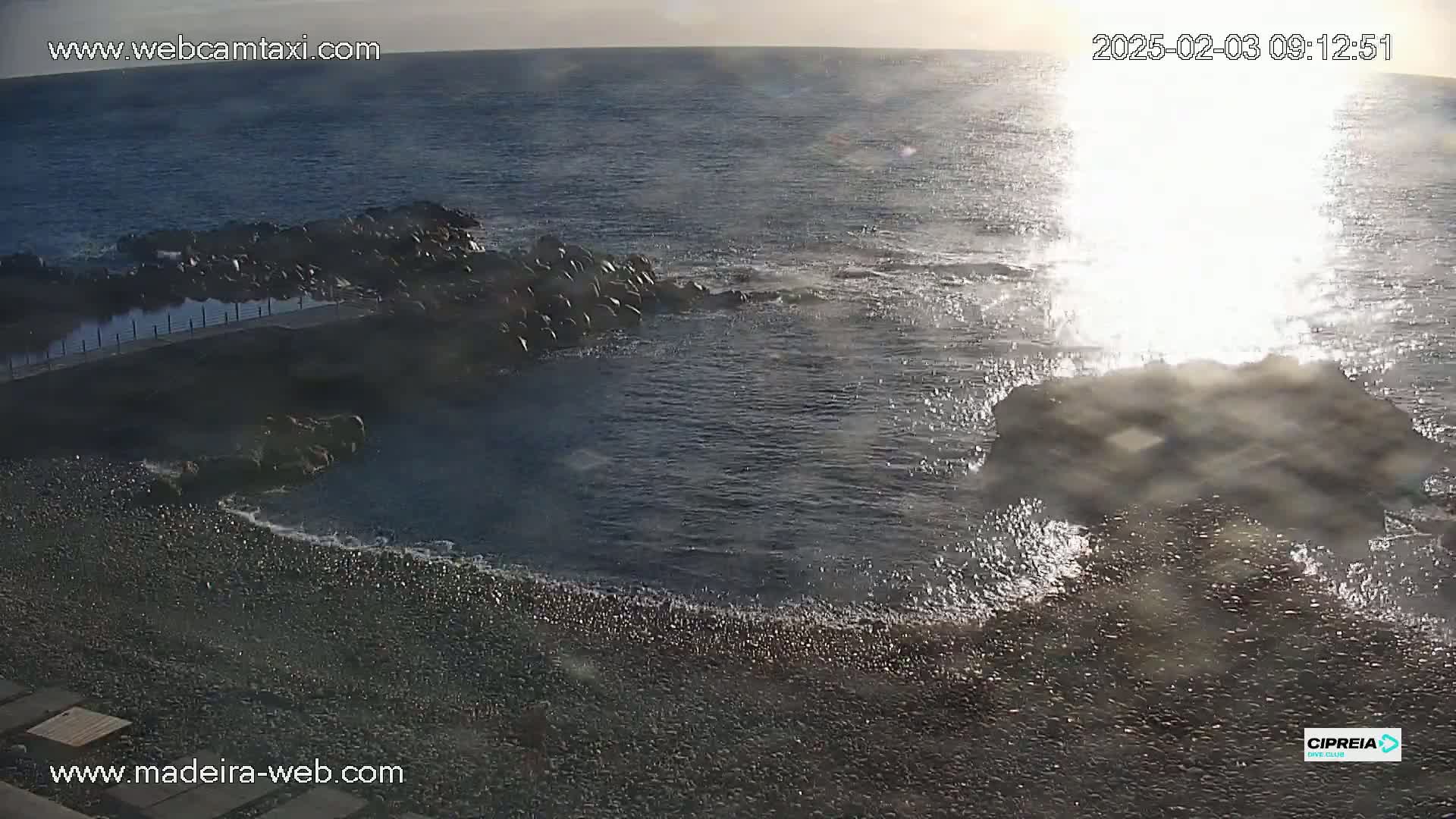 A rocky shoreline with a pebble beach meets a dark blue ocean where waves are crashing against rocks, lit by bright sunlight.