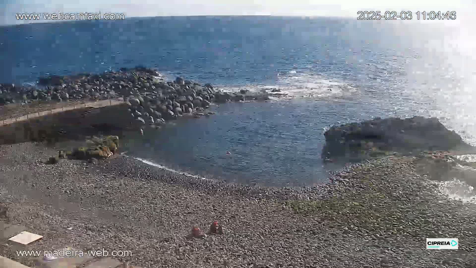 A rocky beach with a calm, partially enclosed body of water, under sunny skies.