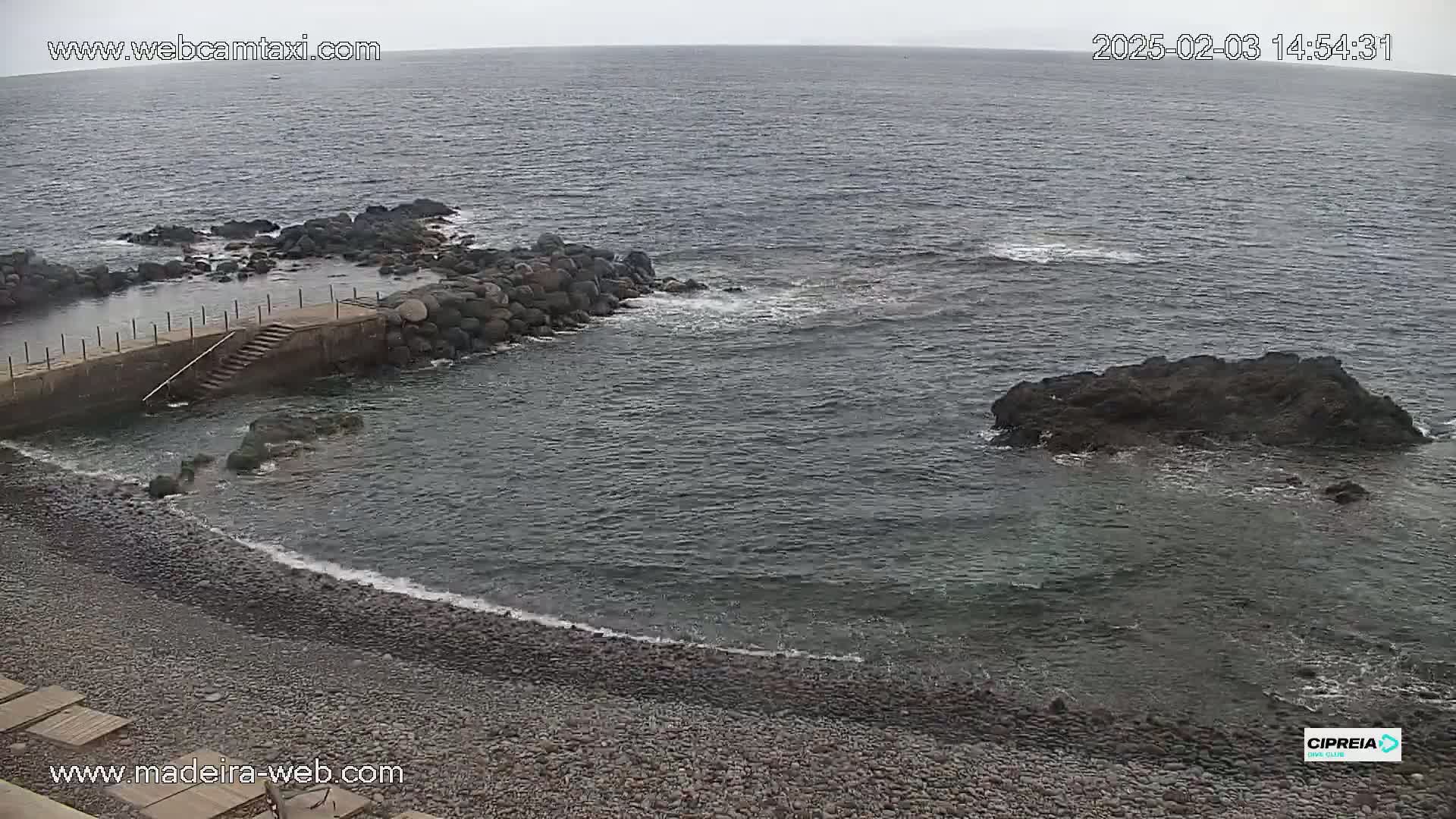 A rocky coastline with a small, stone jetty and a pebble beach is visible under an overcast sky, with gentle waves lapping the shore.