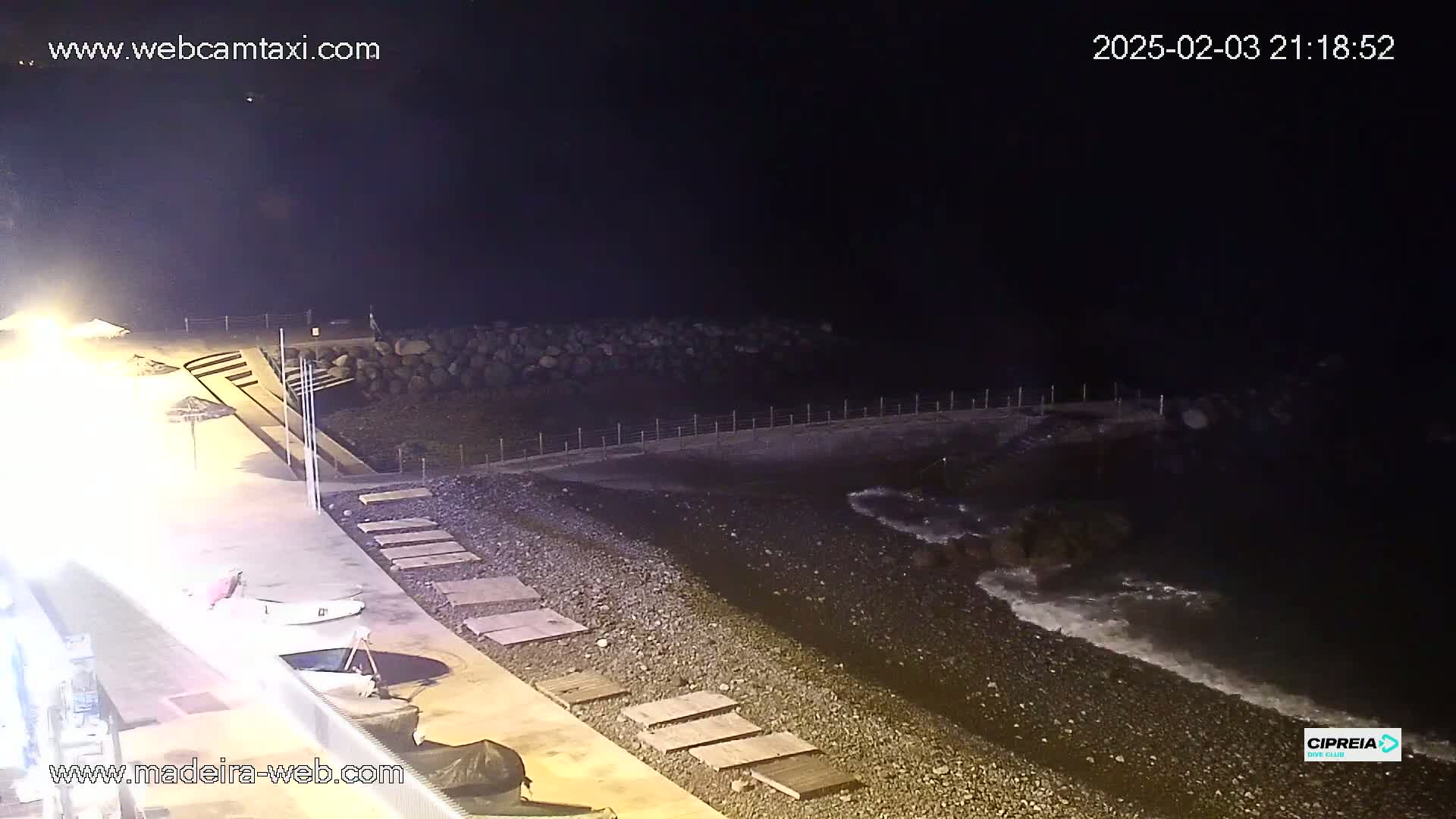 A dark, nighttime beach scene shows a rocky shoreline with gentle waves lapping at the shore, a paved walkway, and some small boats.