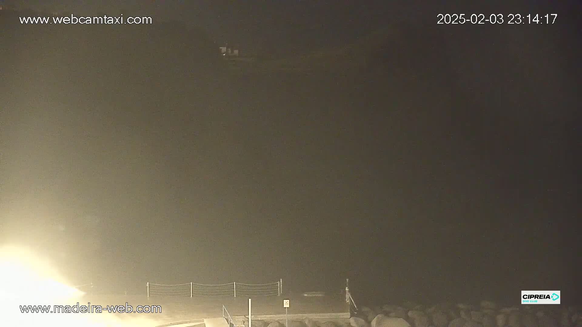 A nighttime, foggy view of a coastal area with a fence and some structures visible in the distance.