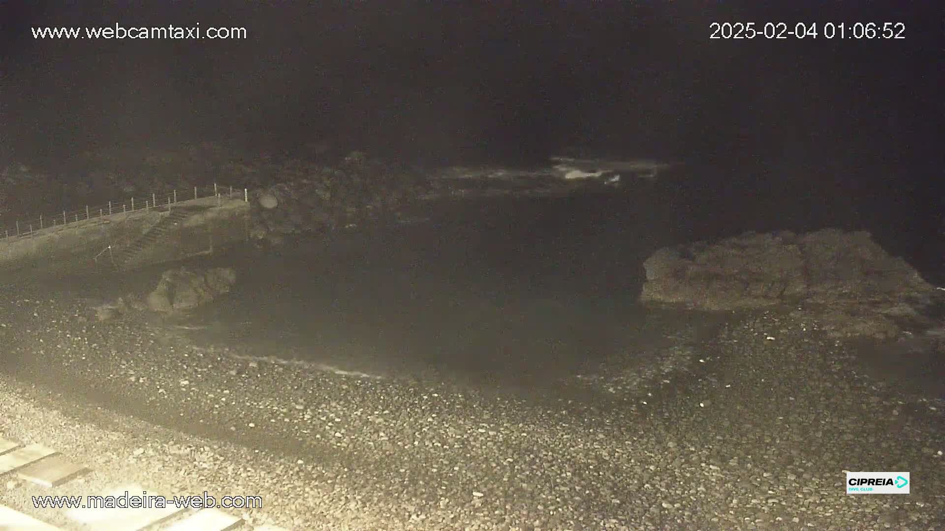 A dark, nighttime view of a rocky beach with calm waves lapping the shore and a set of stairs leading down to the water.