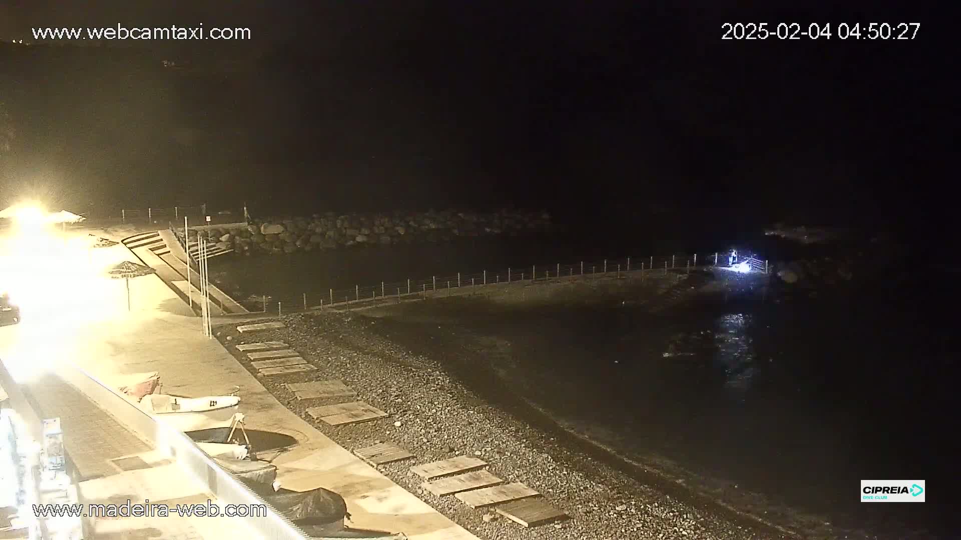 A dark, nighttime view of a pebble beach and calm water, with a few lights visible on the shore and a person standing near the water's edge.