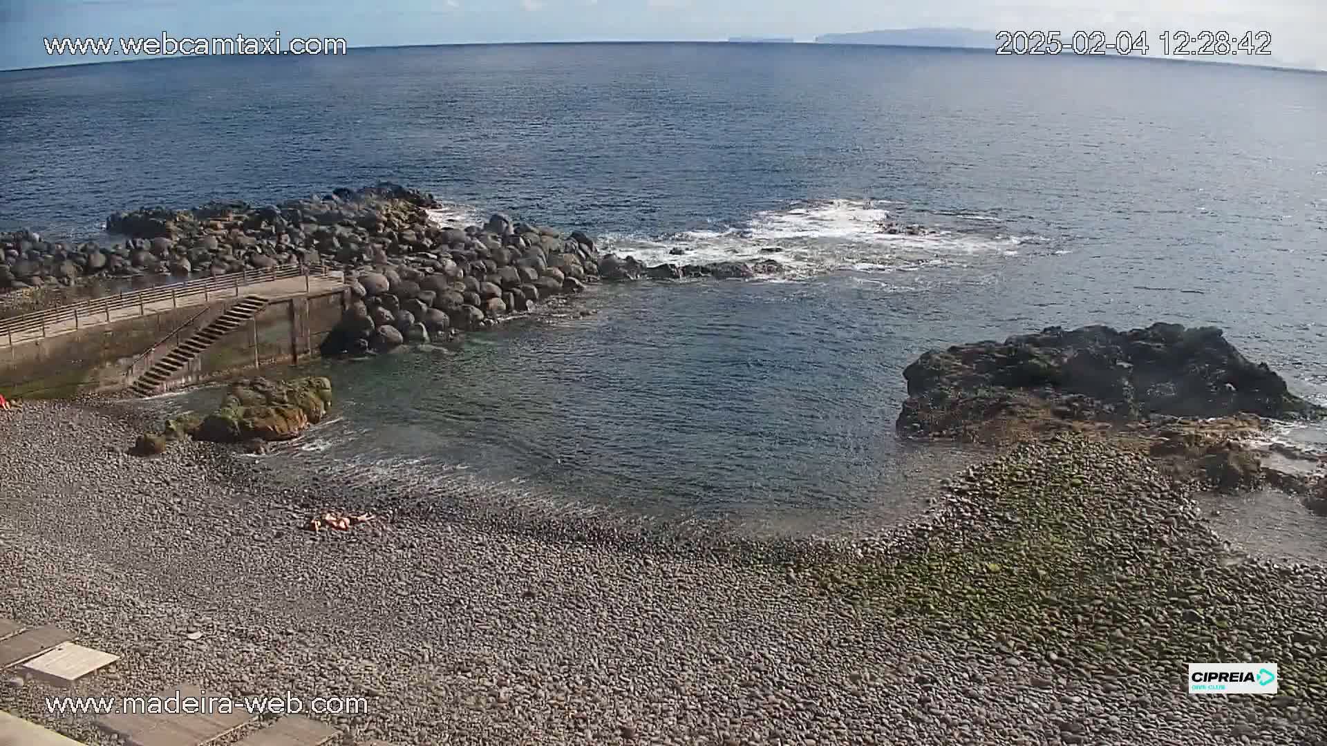 A rocky beach with calm water and two sunbathers is visible on a sunny day.