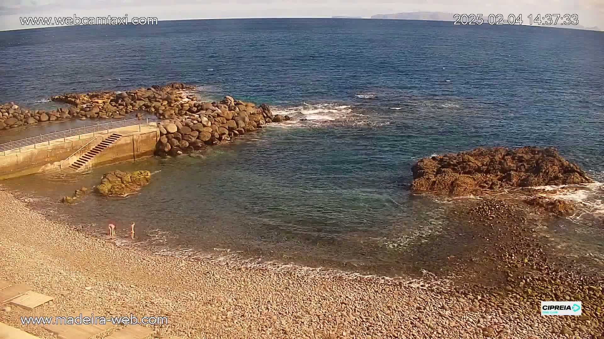 A rocky beach with calm, clear water laps a shoreline with a small man-made harbor and a larger rock formation offshore under a mostly clear sky.