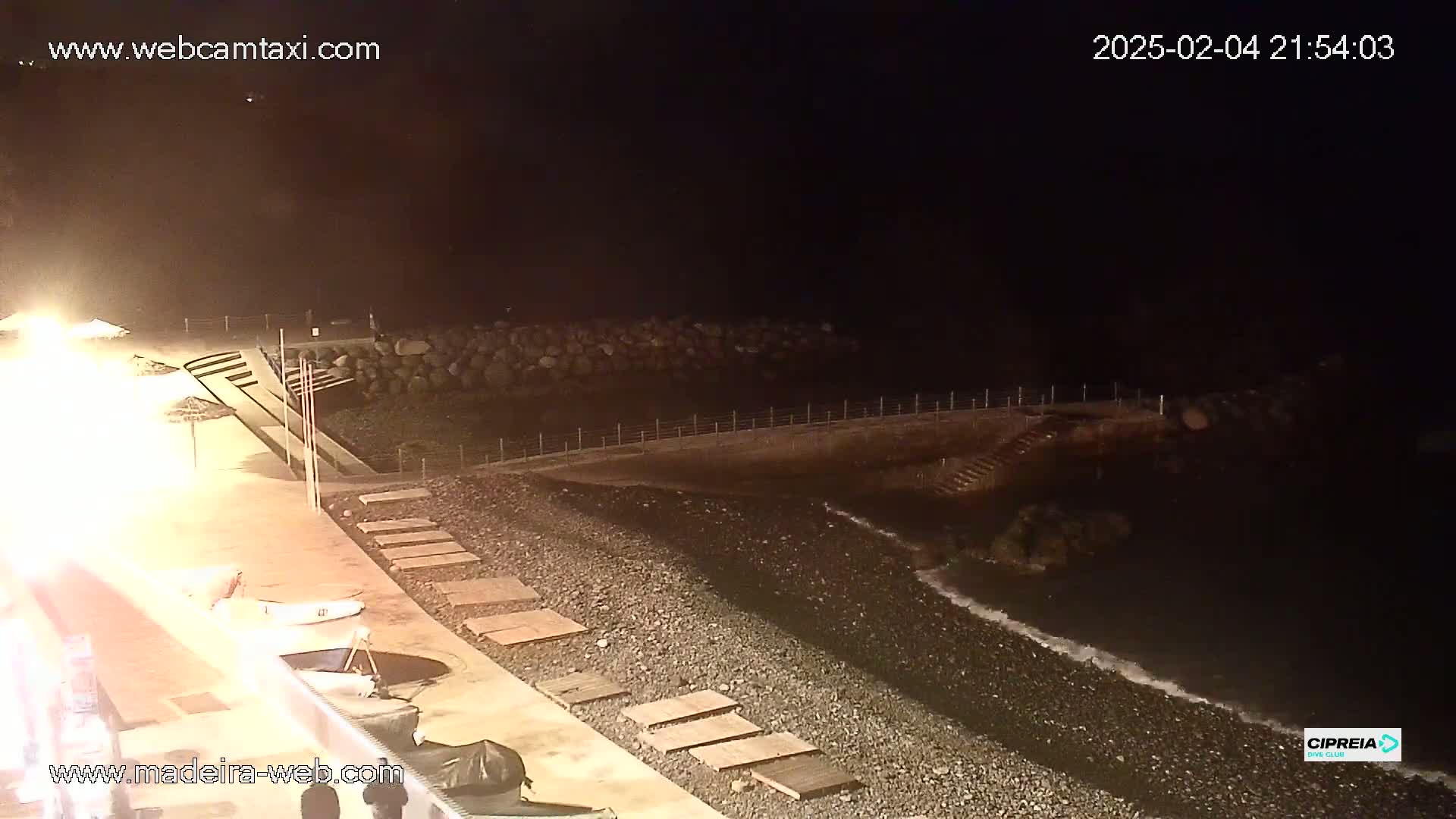 A nighttime view of a dark pebble beach with gentle waves, a low seawall, and steps leading down to the water, under a clear, dark sky.