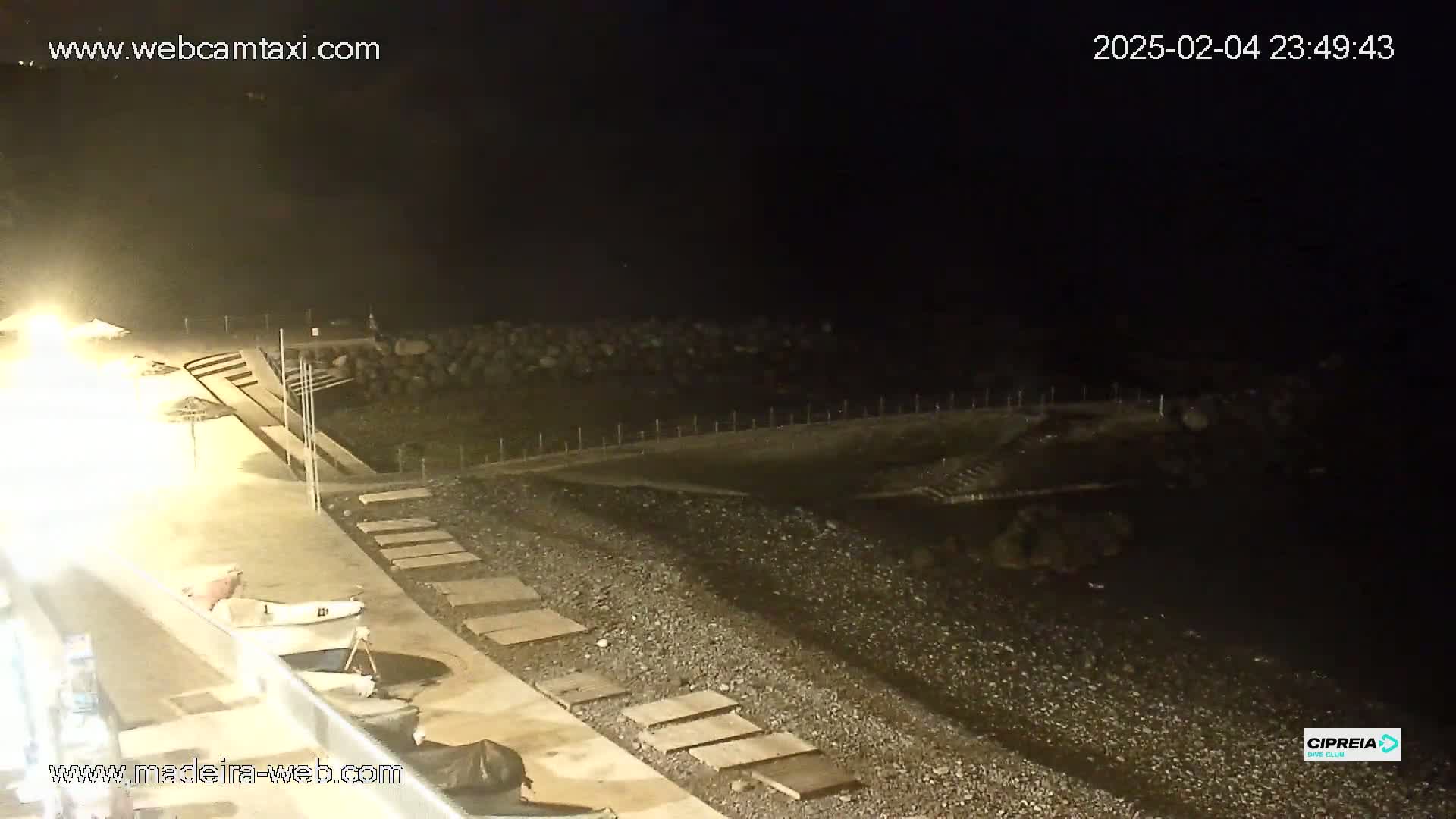 A dark, nighttime view of a pebble beach with steps leading down to the water, a paved area with some small boats, and a partially visible building.