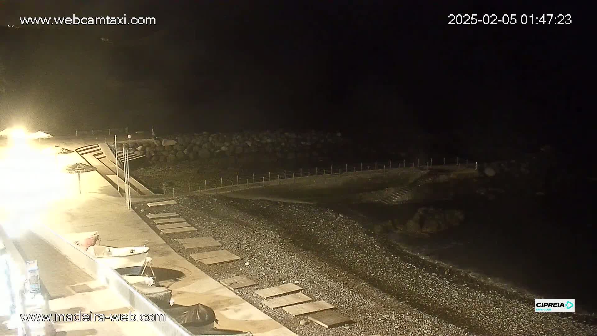 A nighttime view of a pebble beach and paved walkway, with a small boat visible near the shore under dim lighting.
