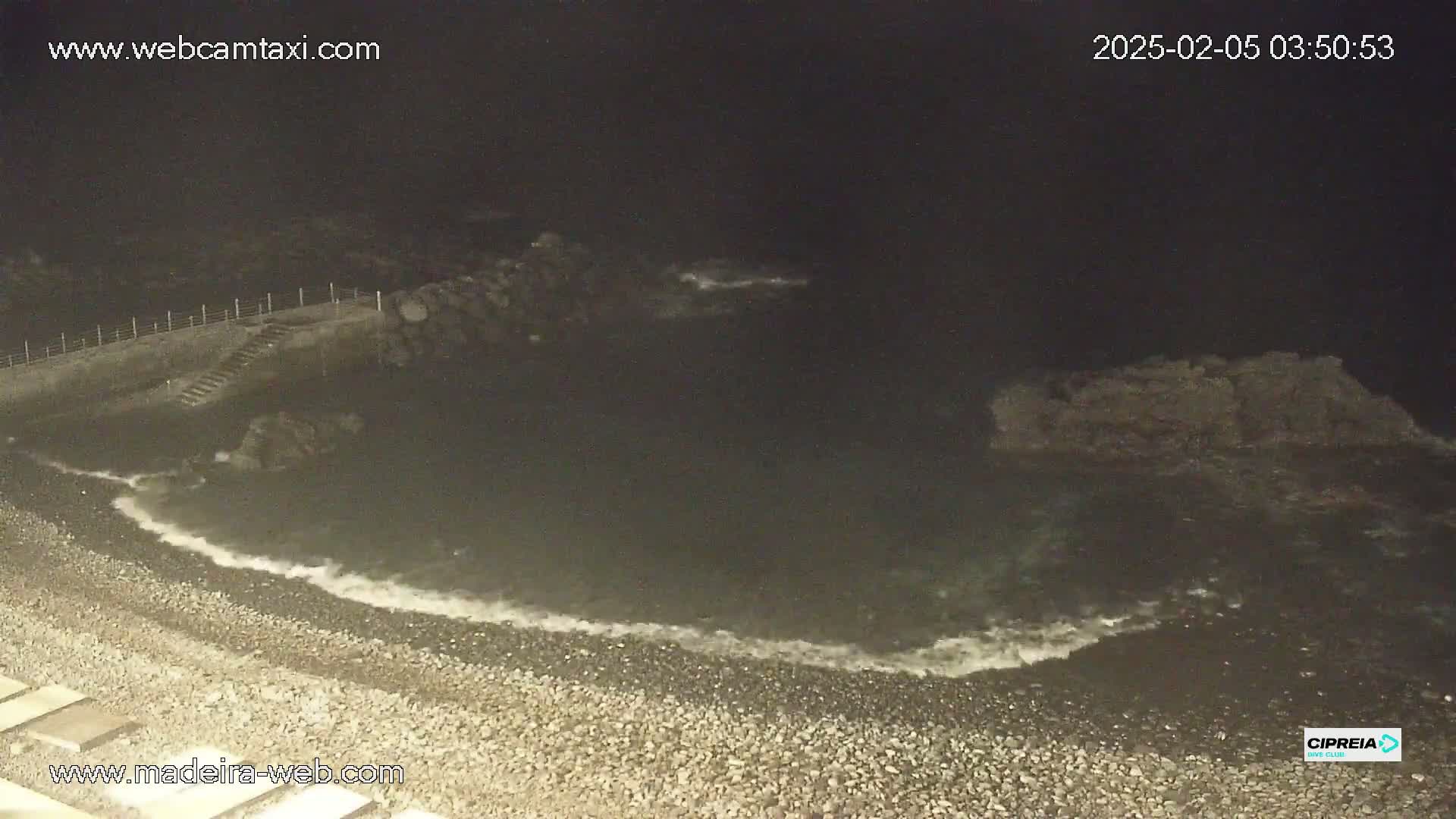 A dark, pebble beach at night with gentle waves lapping the shore near some dark rocks and a set of stairs leading down to the water.