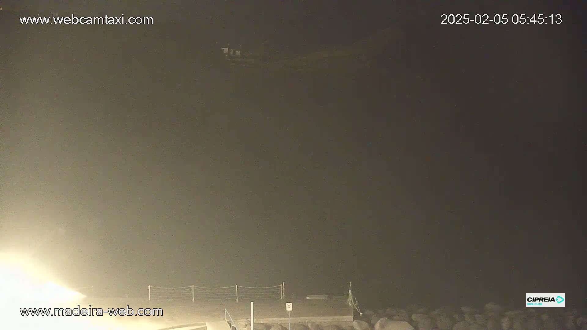 A nighttime, foggy view of a dark body of water, a shoreline with a fence, and some rocks.
