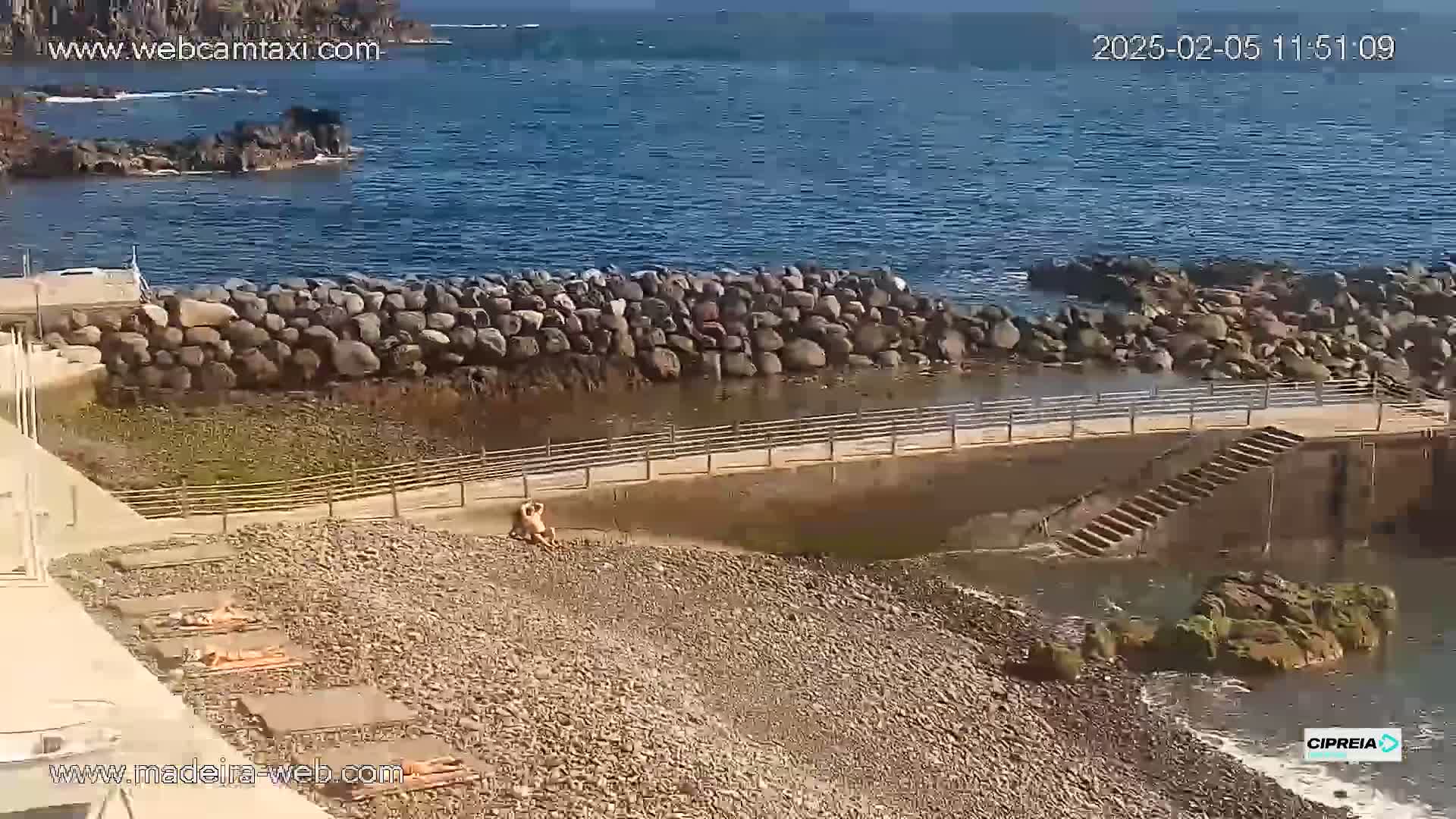 A rocky beach with several people sunbathing is next to a calm ocean under a clear sky.
