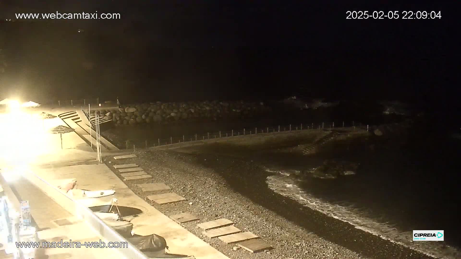 A dark, pebble beach at night with gentle waves lapping the shore, illuminated by artificial lights from a nearby building.