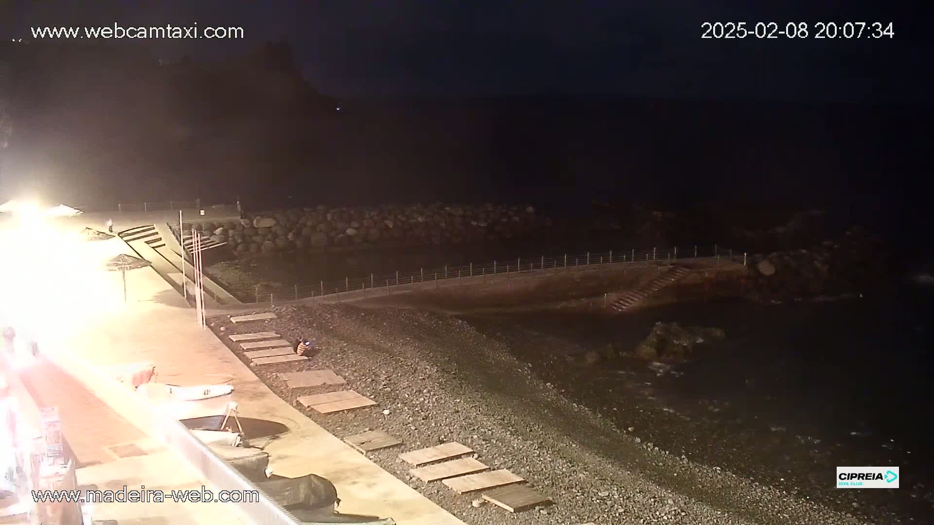 A dark, nighttime view of a pebble beach and a rocky shoreline, illuminated by artificial lights.