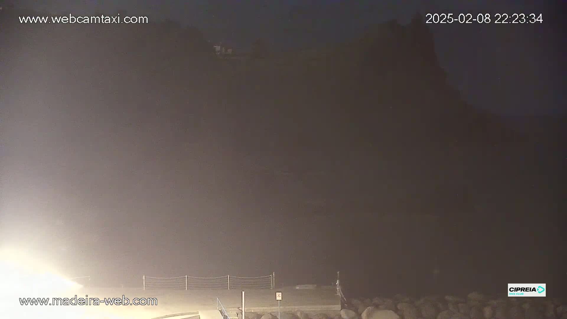 A dark, foggy night scene shows a dimly lit shoreline with a rocky area in the foreground and a dark cliff face in the background.
