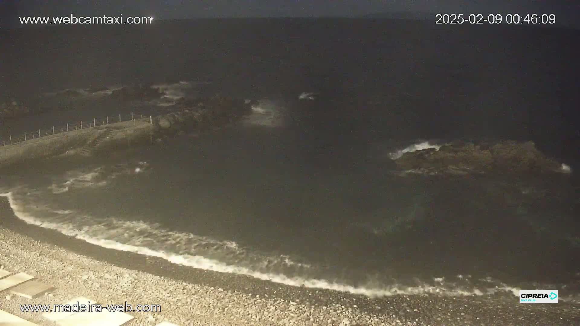 A dark, nighttime beach scene shows gentle waves lapping at a pebble shore near a low wall and some rocks jutting out into the water.