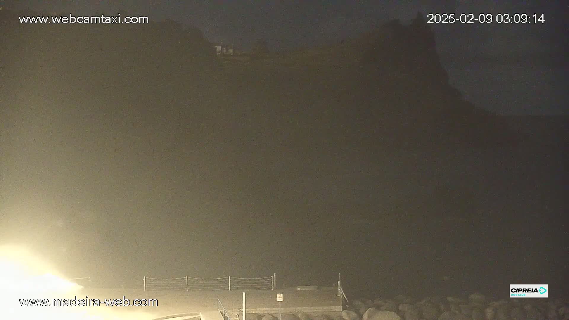 A nighttime, hazy view shows a dark cliff face with a few lights visible in the distance, and a low-lying, dimly lit area at the bottom.