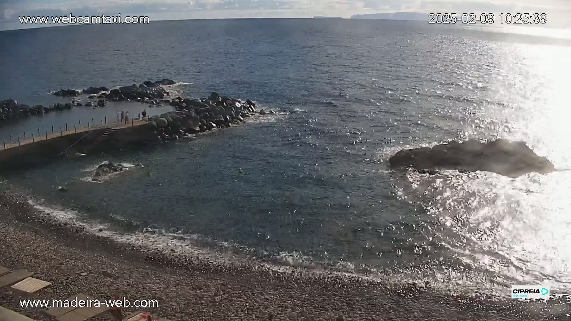 A rocky shoreline with a small, calm swimming area, a few people on a pier, and waves crashing against rocks in the ocean under partly cloudy skies.