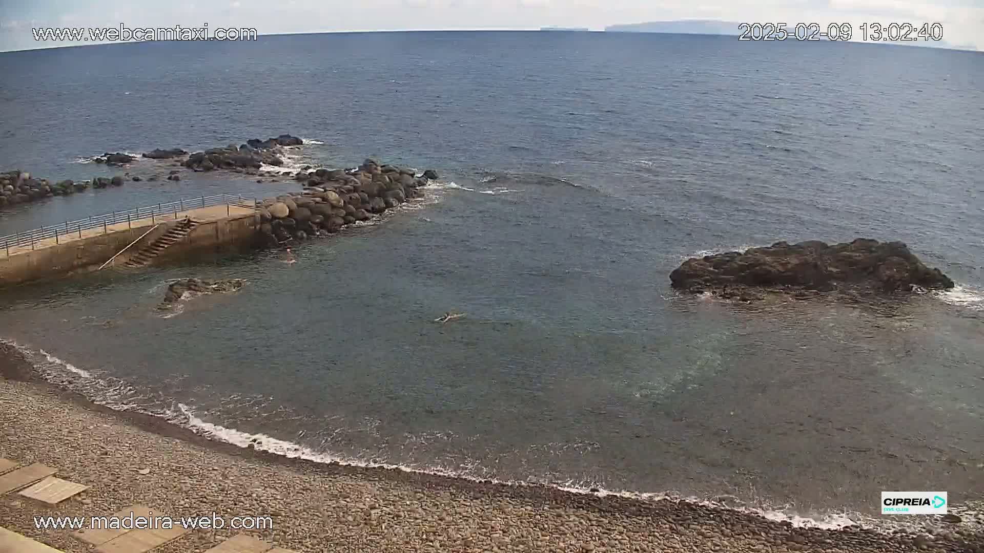 A rocky, pebble beach with calm, clear water, a small concrete pier with stairs leading into the ocean, and a person swimming in the sea under a partly cloudy sky.