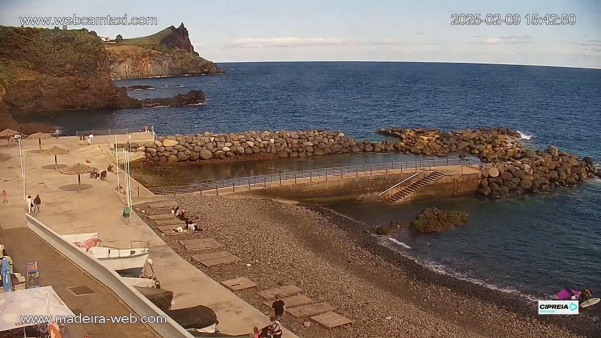 A rocky beach with a small harbor and calm ocean under a mostly clear sky, several people are scattered on the beach and near the water.