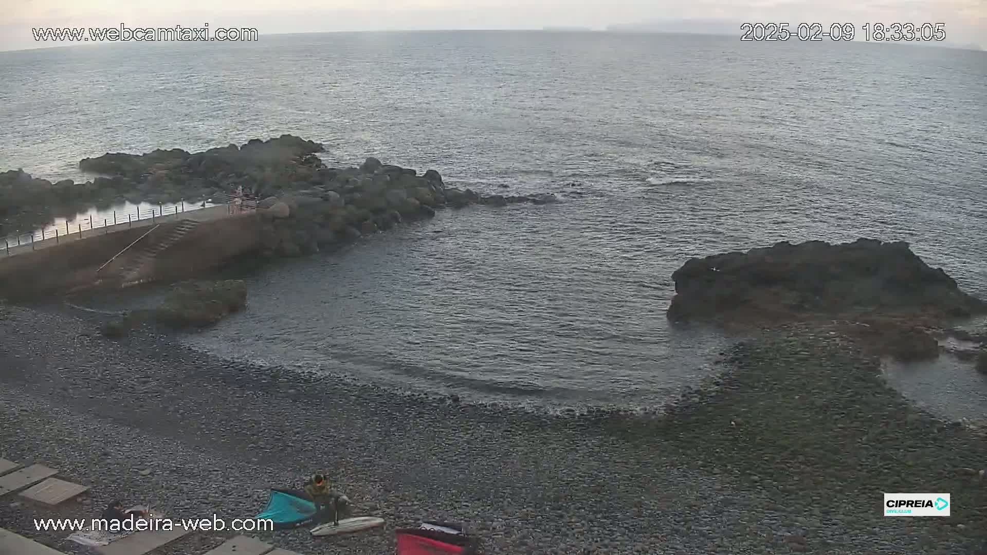 A rocky, pebble beach meets a calm ocean under an overcast sky, with a few people visible near a small stone jetty.