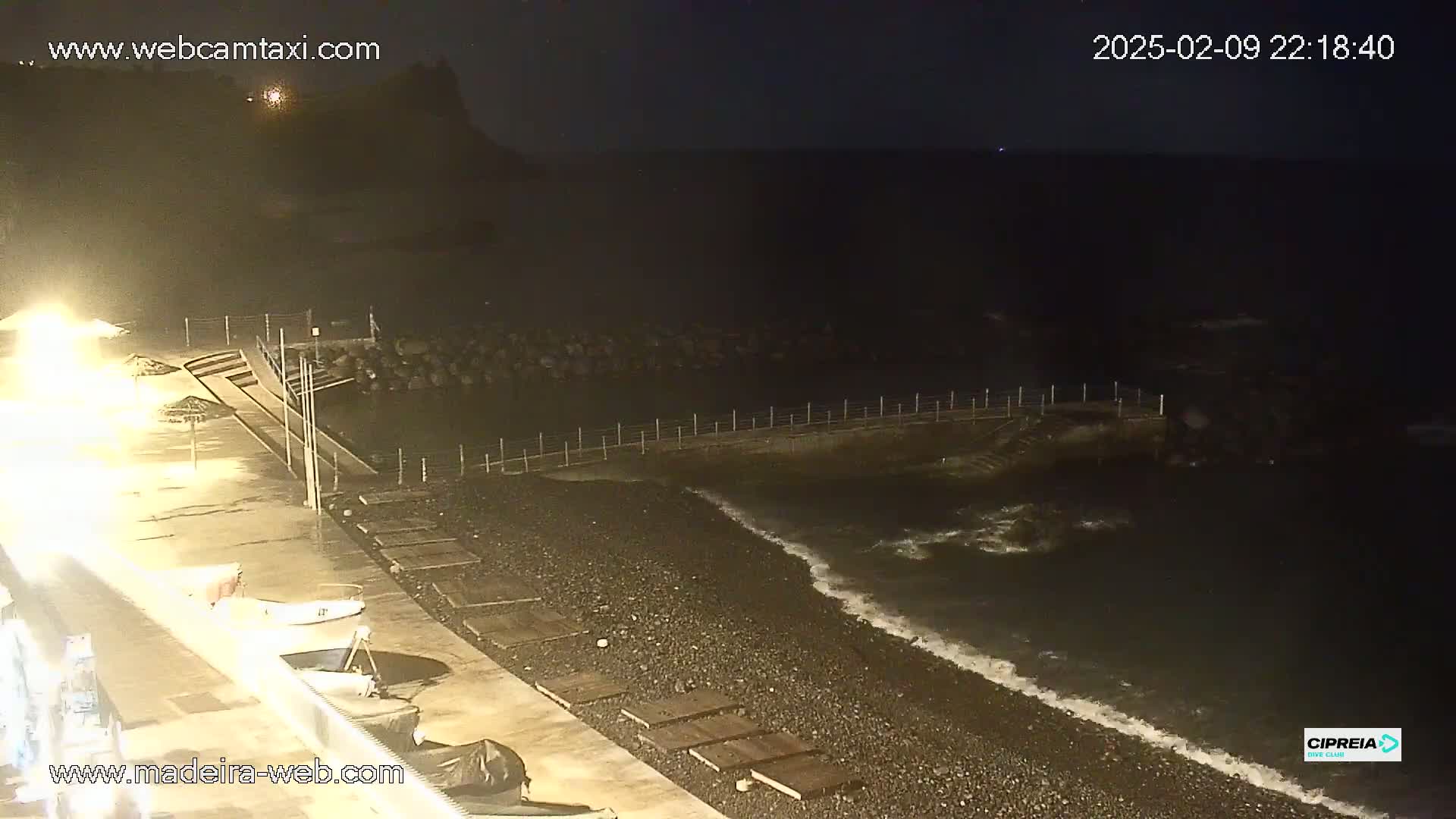 At night, a dark, rocky beach is gently lapped by waves under a clear, starlit sky; a small boat is moored nearby.