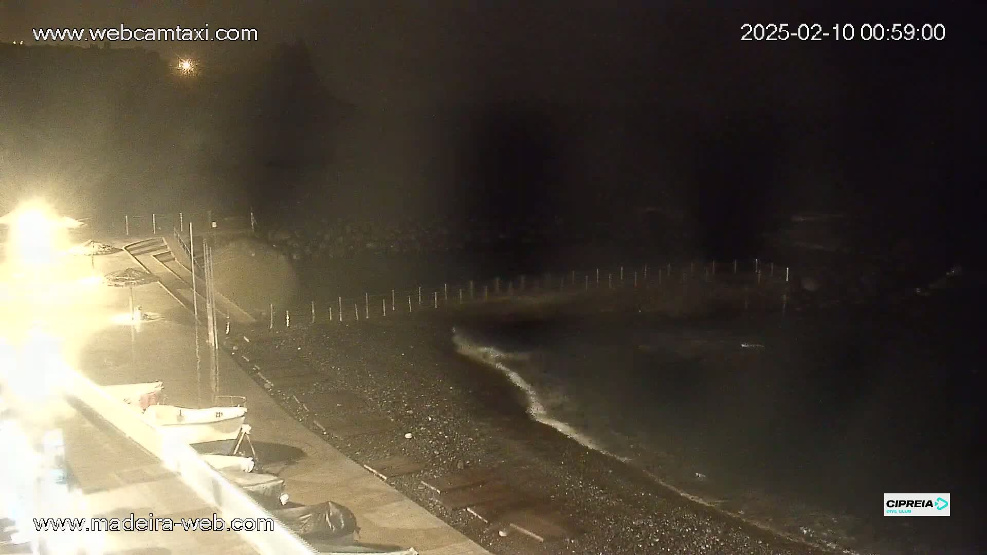 A nighttime view of a dark, pebble beach with gentle waves lapping the shore, illuminated by artificial lights, and a few small boats moored nearby.