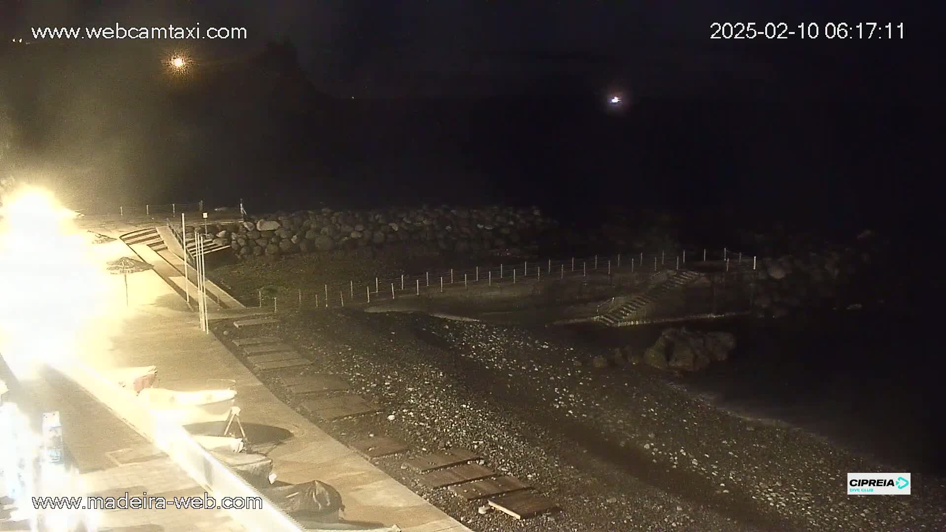 A nighttime view of a dark pebble beach with a boardwalk, steps leading down to the water, and a rocky barrier separating the land from the sea.