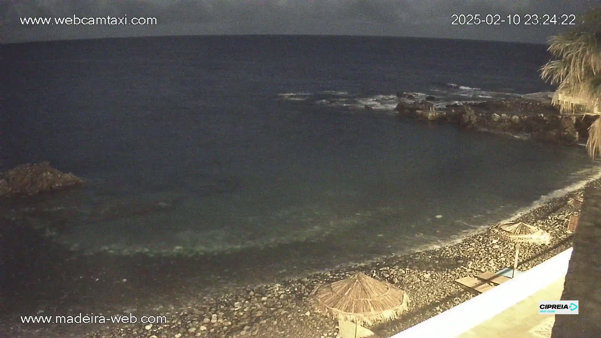 A dark, rocky beach at night with calm, dark water and a few thatched umbrellas on the shore under an overcast sky.