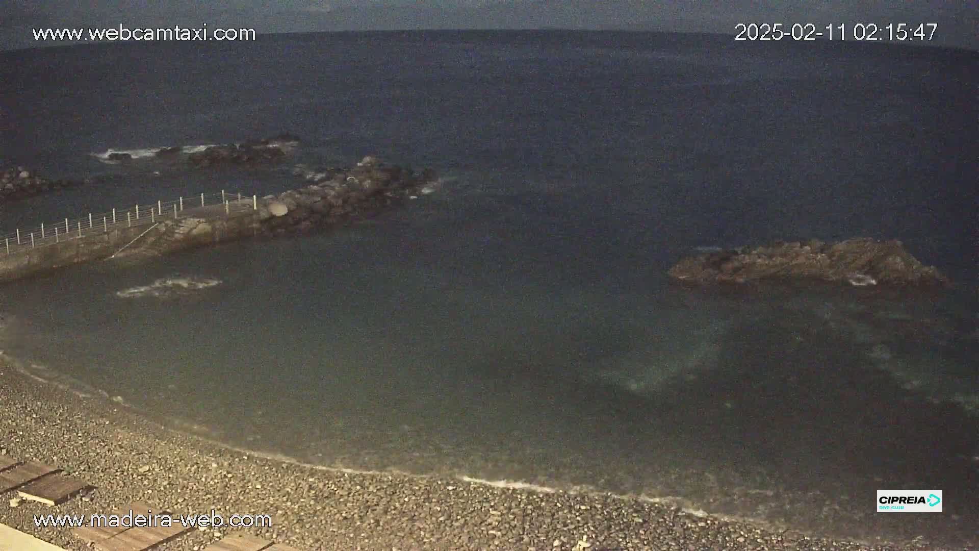 A dark, pebble beach meets a calm, dark-colored sea at night, with a small jetty and rocky outcroppings visible in the water.