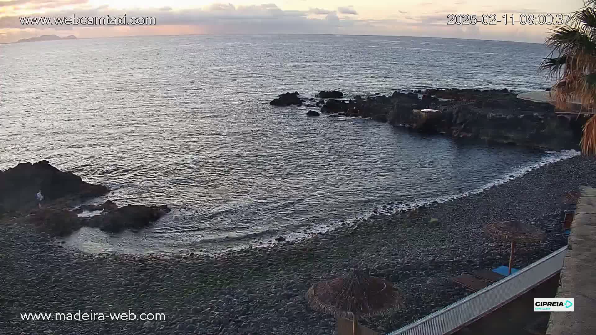 A rocky, dark-pebble beach meets a calm, dark-grey ocean under a partly cloudy, dusky sky.