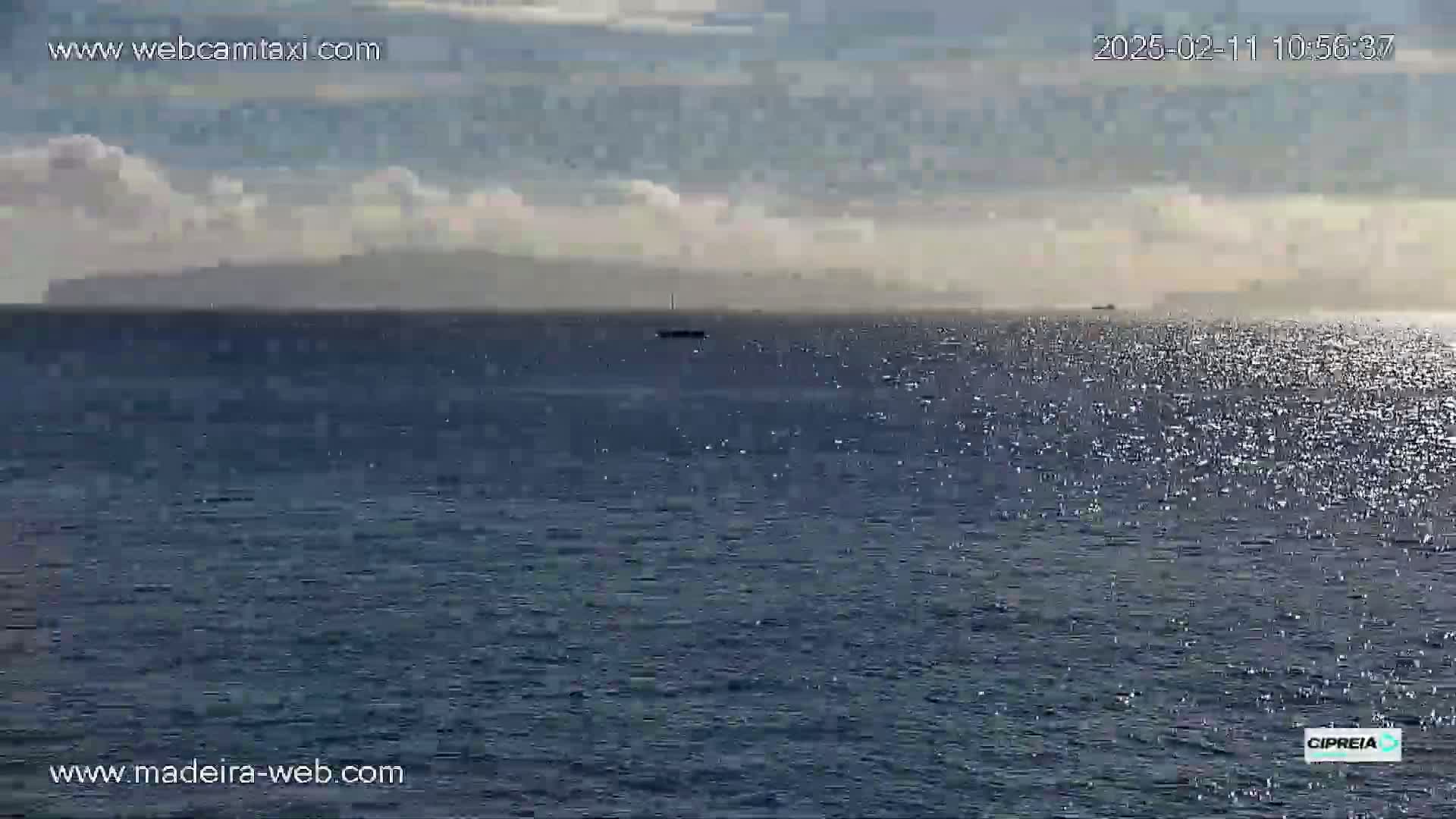 A partly cloudy day shows a calm ocean with a distant landmass and a small vessel on the horizon.