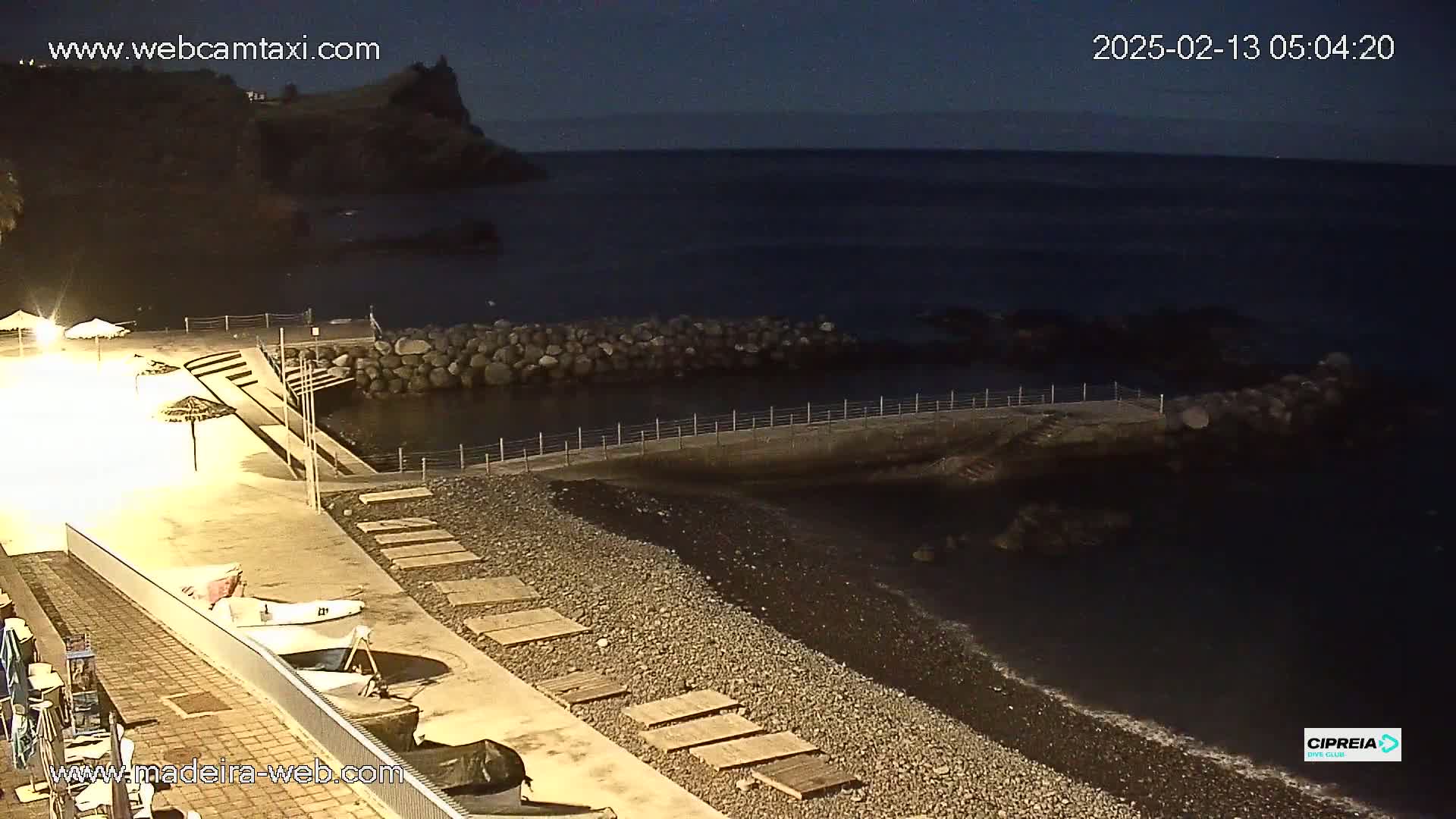 A dark, nighttime beach scene shows a pebble shore, a small harbor with a low stone wall, and a paved area with some beach umbrellas and small boats.