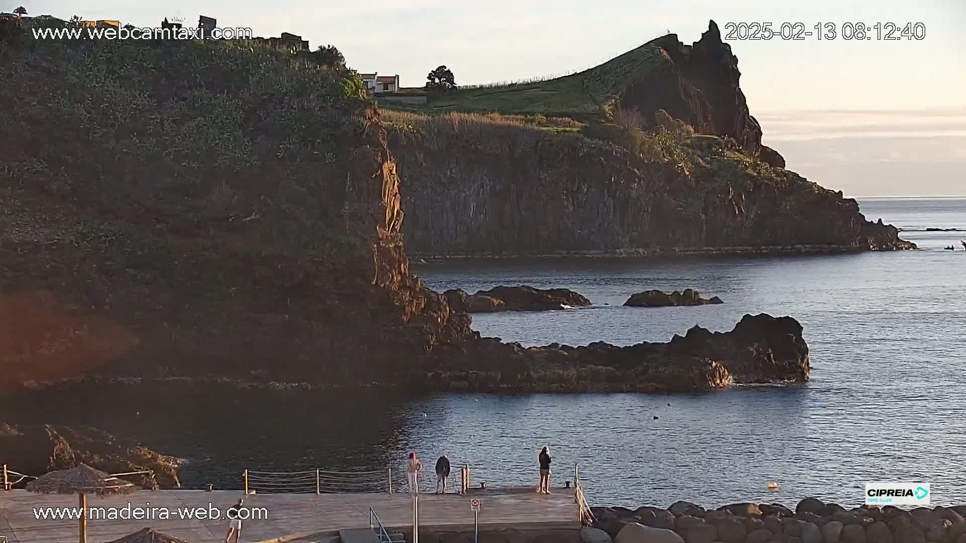 A rocky, dark-colored coastline with a small dock and three people standing on it, under a calm, clear sky at sunrise or sunset.
