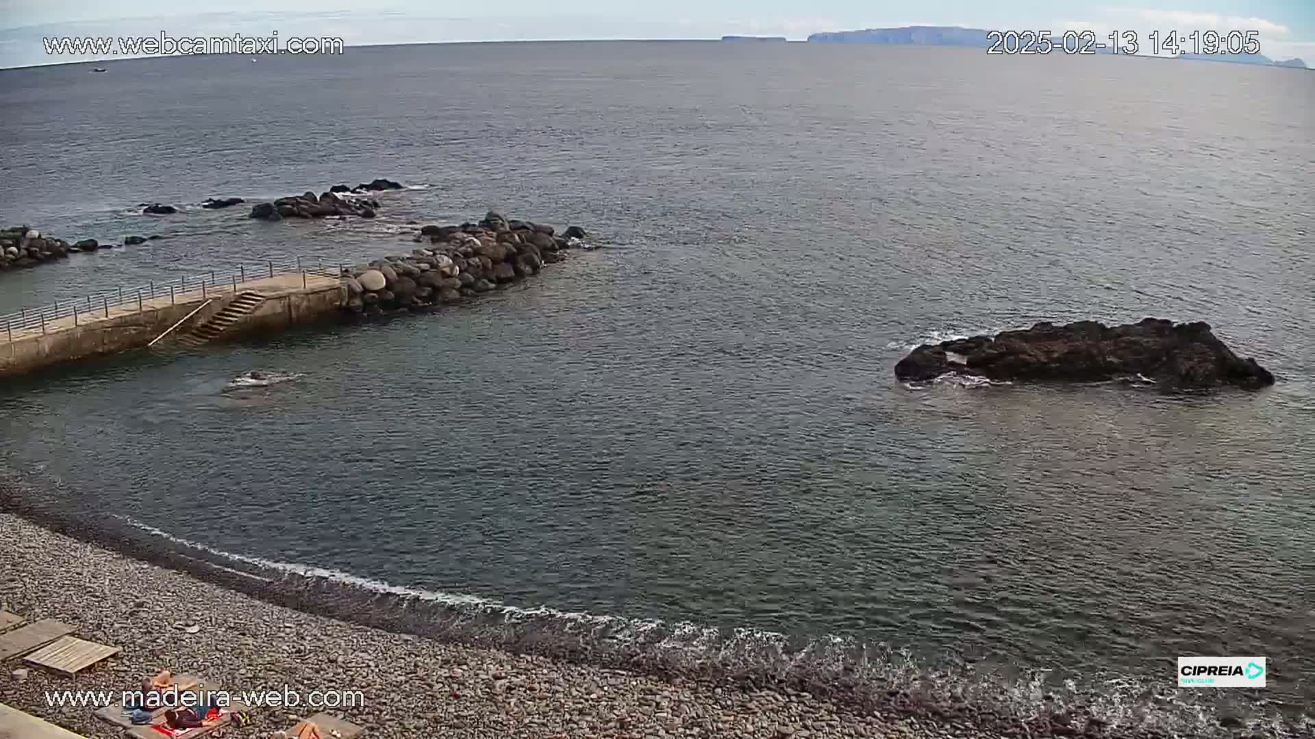 A rocky beach with calm, dark water laps gently at the shore, near a small stone jetty, with a distant landmass visible on the horizon under an overcast sky.