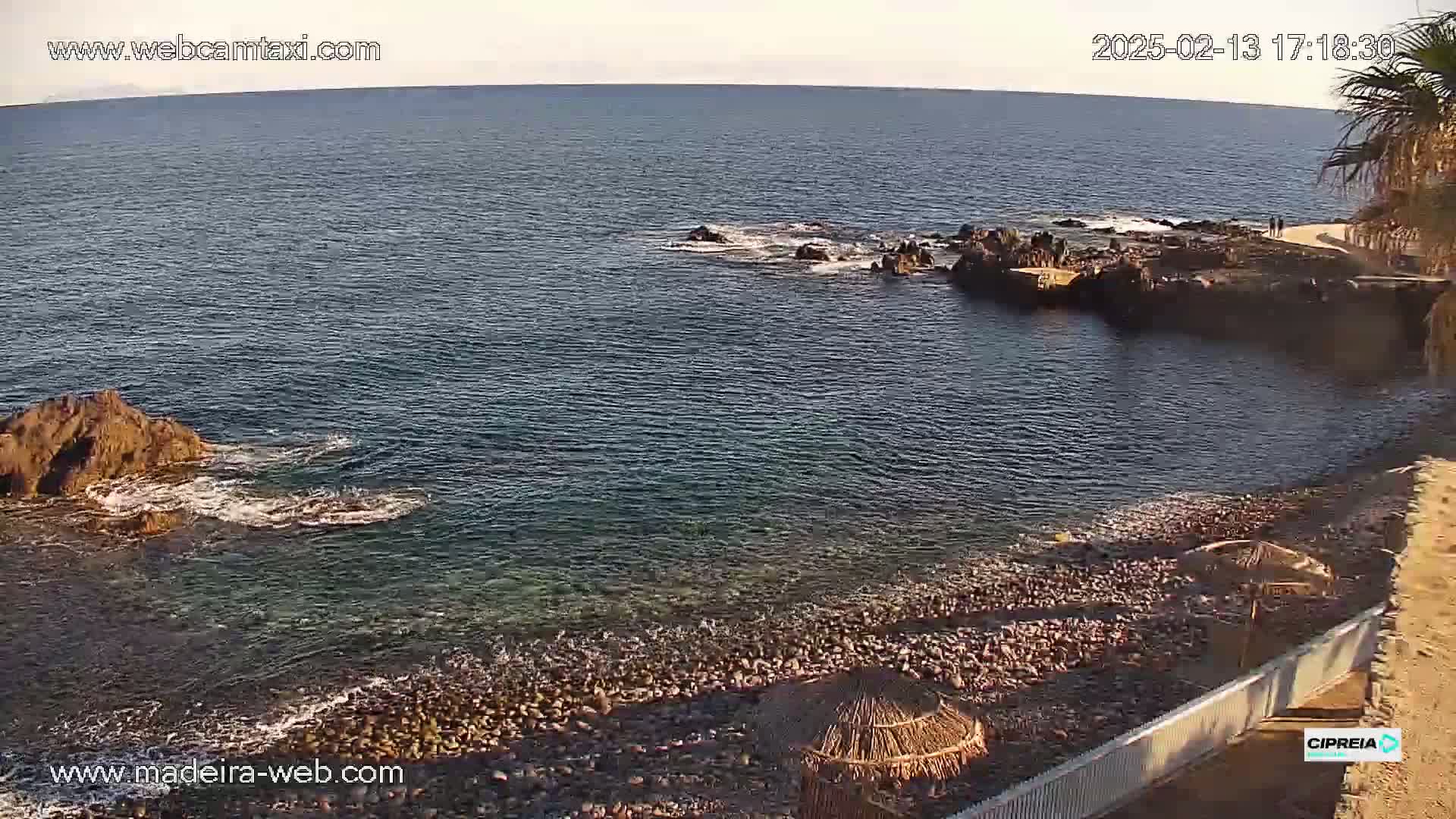 A rocky shoreline with calm, clear water under a mostly clear sky, features a pebble beach with a few beach umbrellas.
