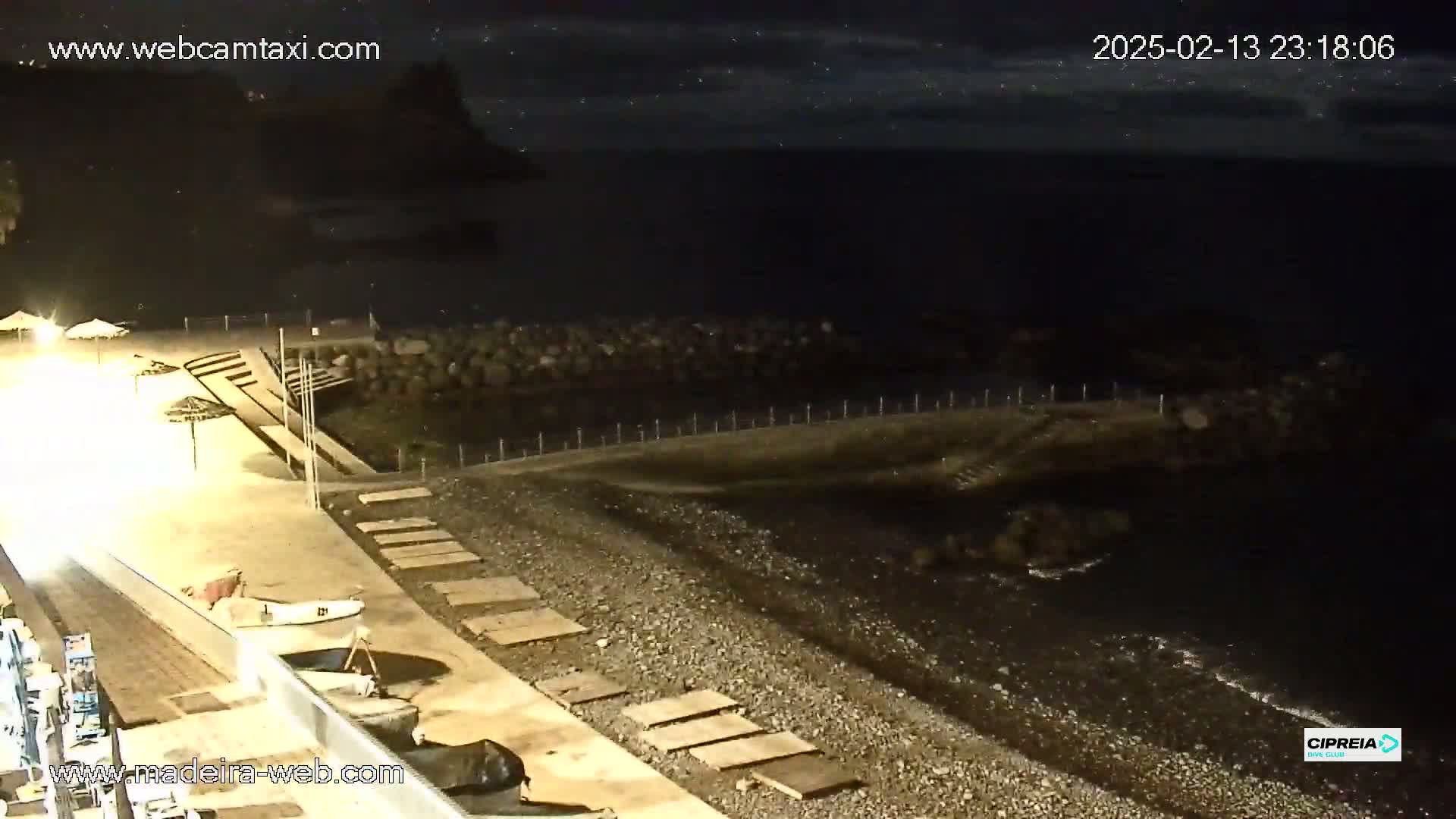 A nighttime view of a pebble beach with a calm, dark ocean, a low seawall, and several closed beach umbrellas.
